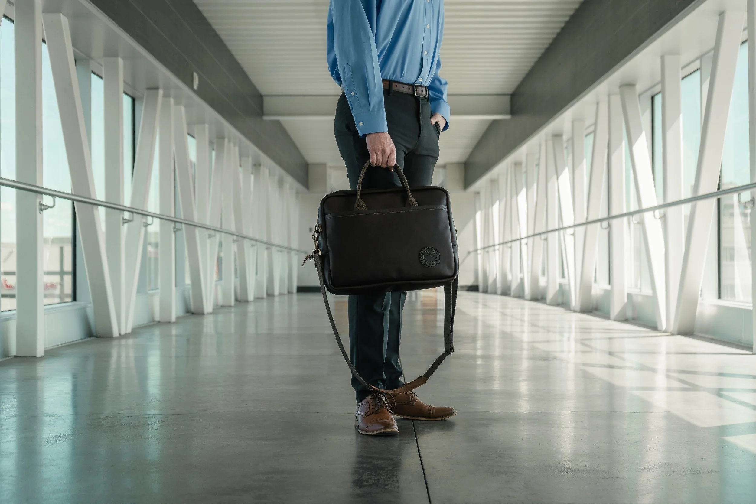 Person in business casual attire holding a black laptop bag standing in an airport terminal with large windows and modern architecture.