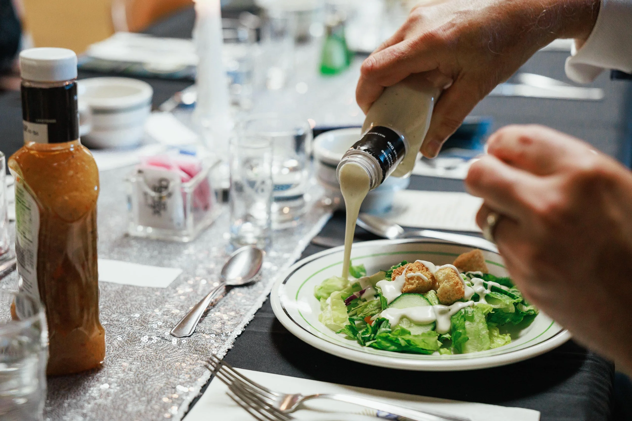 A person pouring salad dressing onto a plate of salad with lettuce, cucumbers, croutons, and dressing, on a table set for a meal.