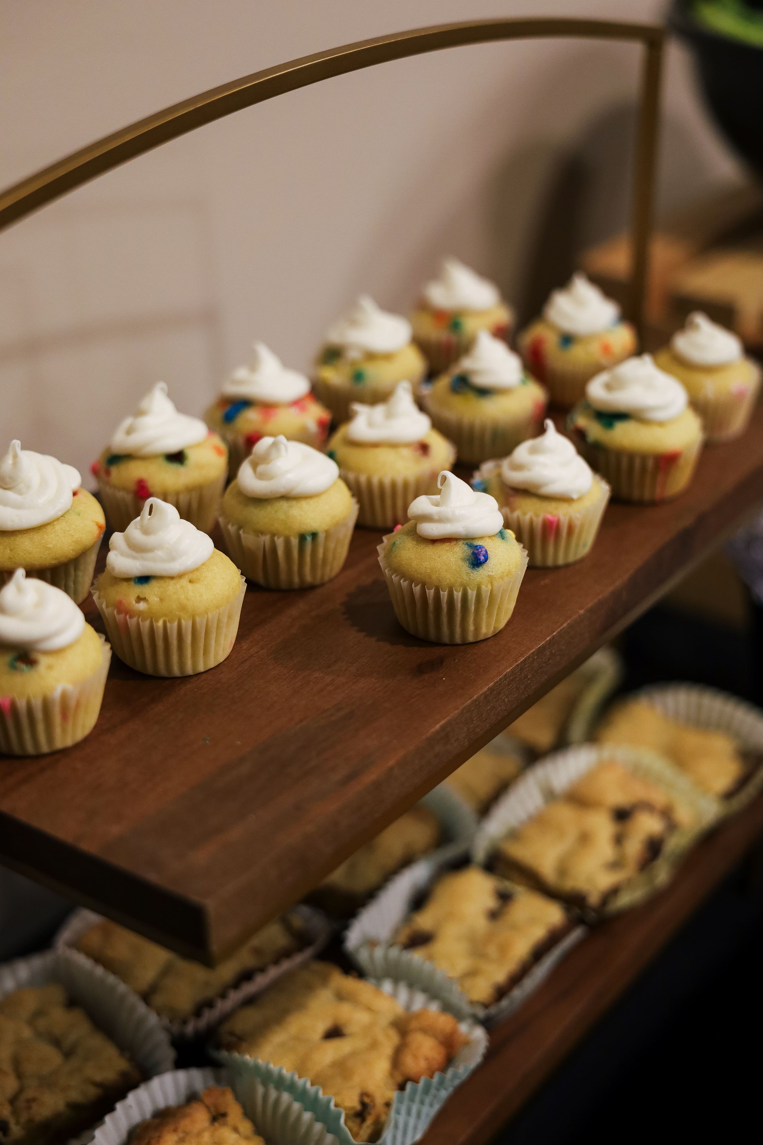 A wooden dessert stand holding cupcakes with white frosting and colorful sprinkles, and a tray of assorted cookies below.