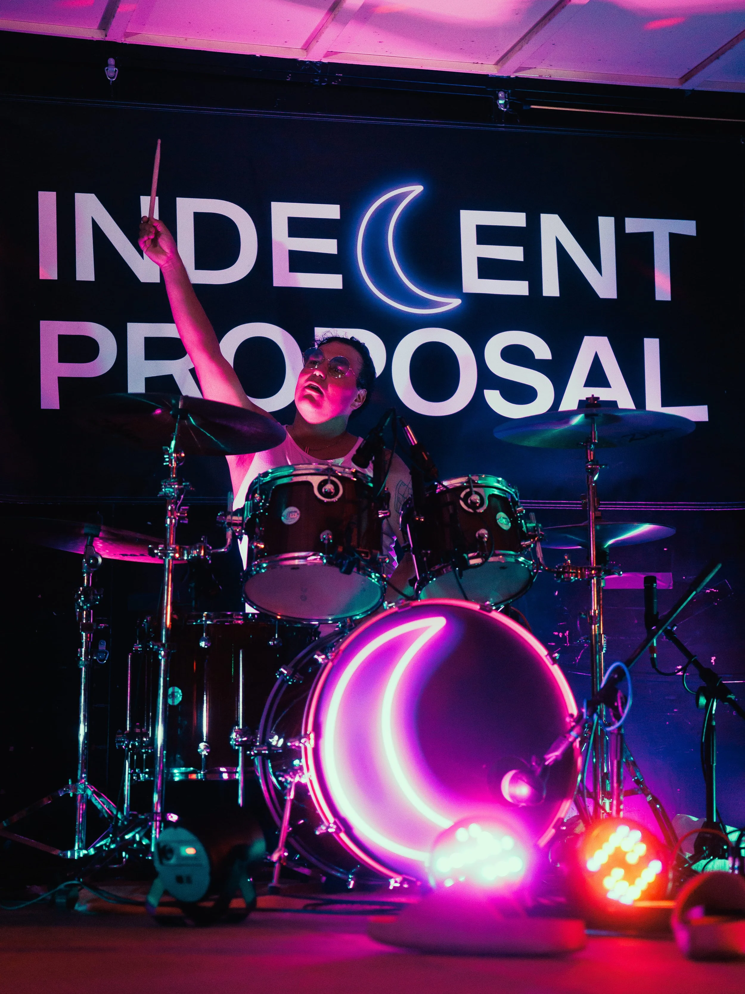 A person playing a drum set on stage with a neon sign behind that reads 'Indecent Proposal' with a crescent moon symbol. The stage lights are pink and purple, casting colorful reflections on the drums.