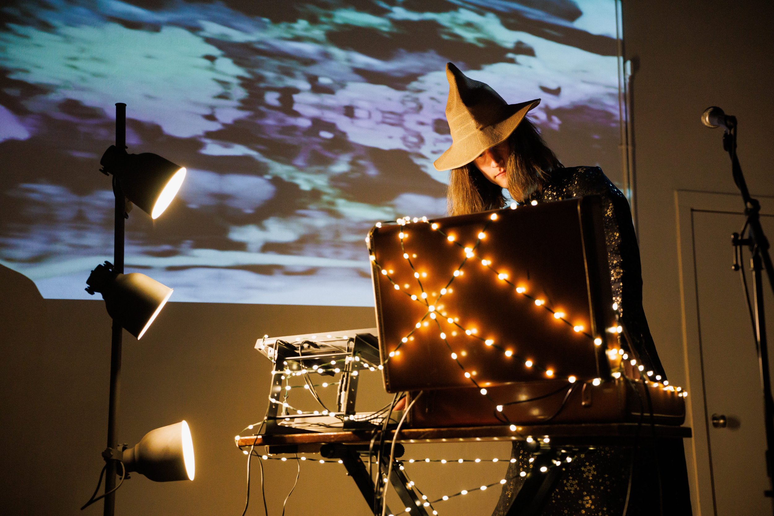 A person wearing a witch hat performs on a keyboard adorned with string lights, with a backdrop of a projected image of water and waves, and stage lights illuminating the scene.
