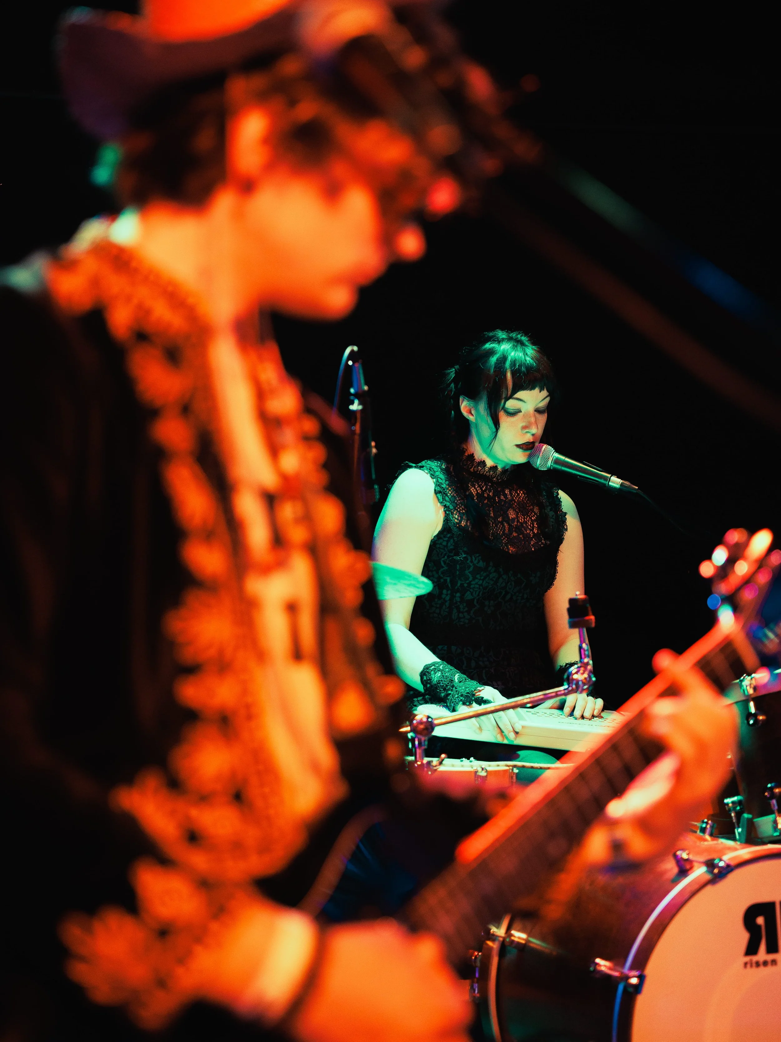 A musician playing a keyboard or synthesizer, dressed in a black lace top, with short black hair and dark lipstick, under colorful stage lighting.
