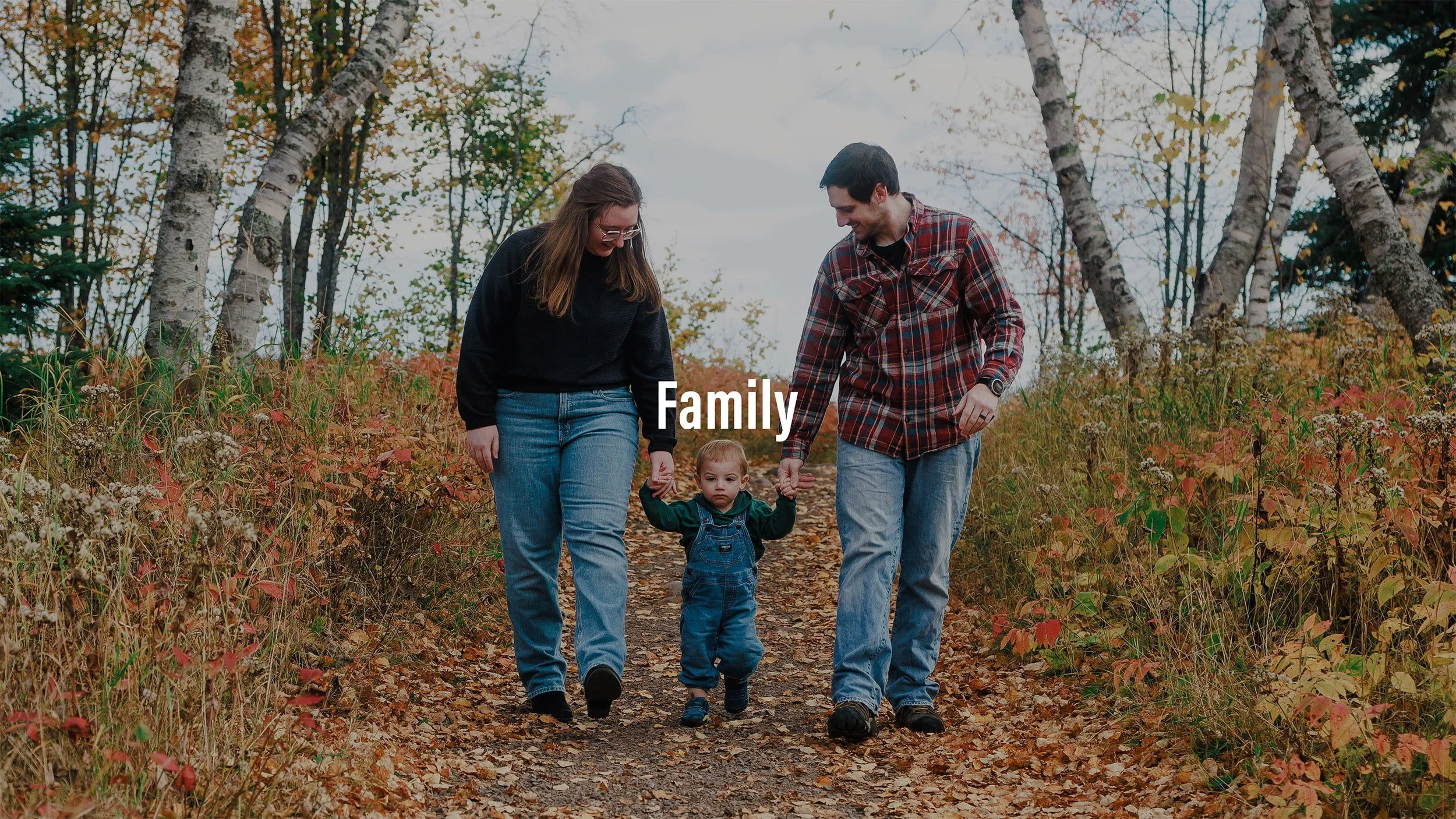 Family walking on a trail in a fall forest, holding their child's hands, surrounded by trees with orange and yellow leaves.
