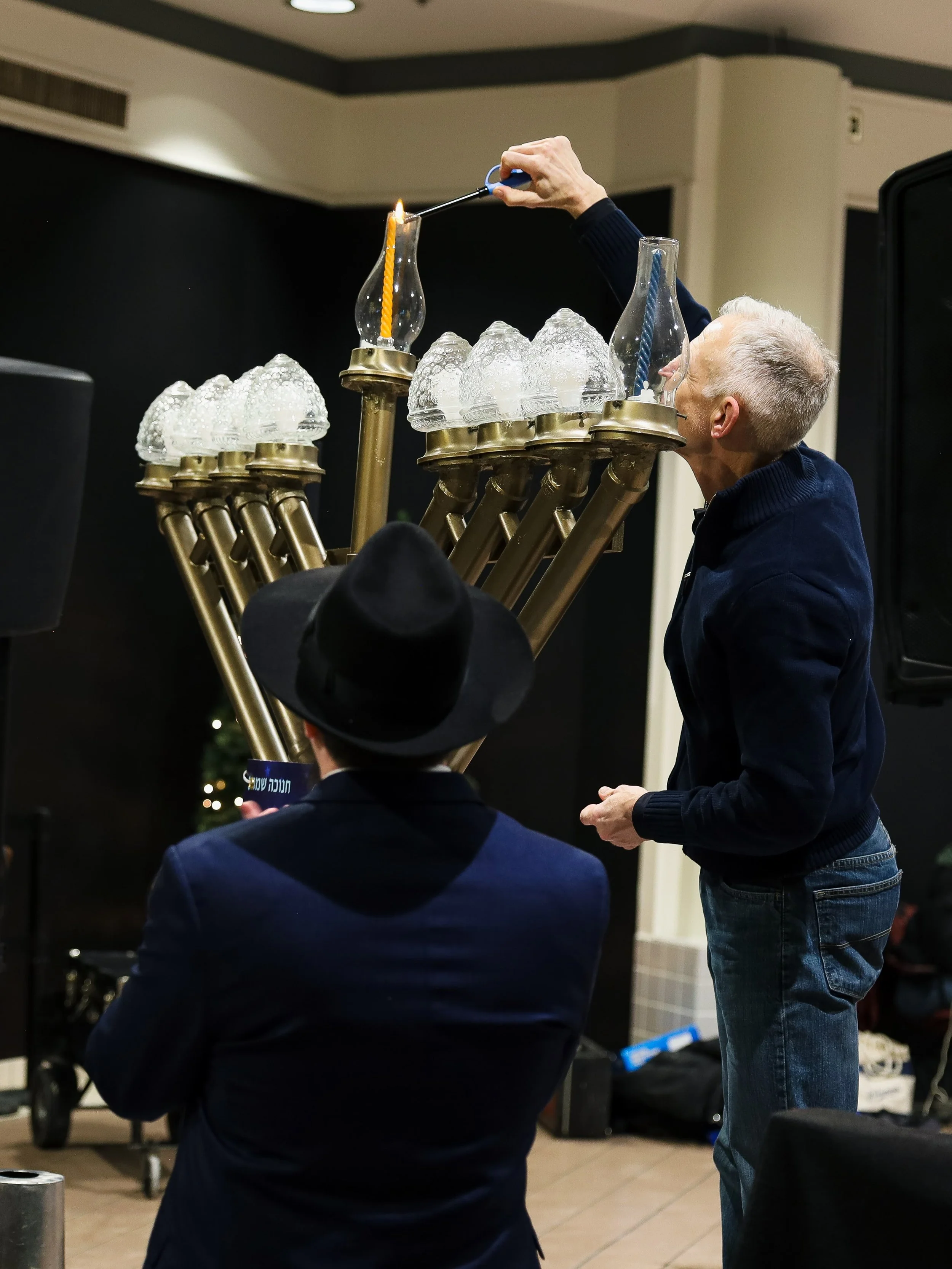 A man lighting a candle on a large menorah at an indoor event, with another person watching.