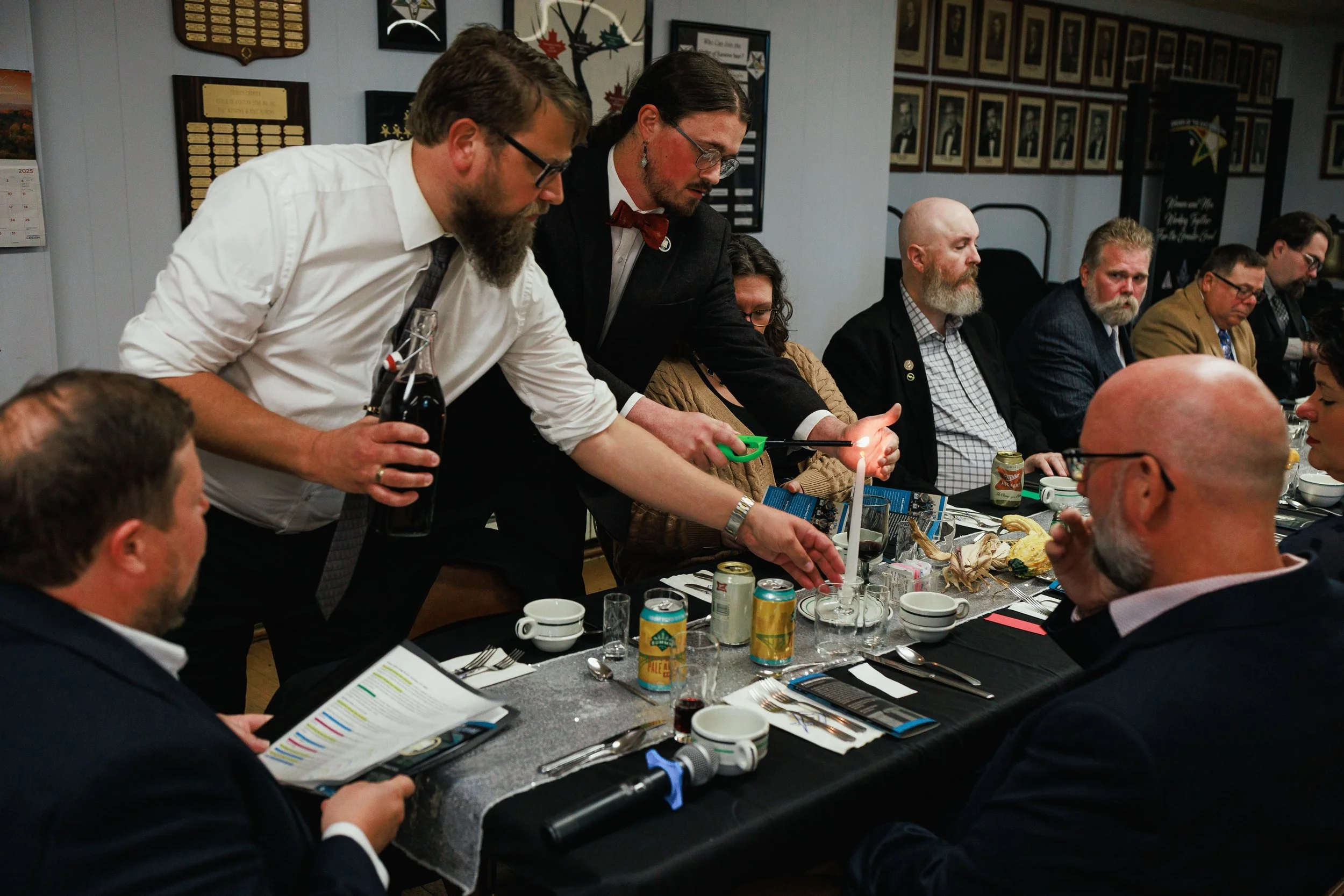 A group of people dressed in formal attire gathered around a table during a meeting or celebration, with two men in the center lighting a candle.