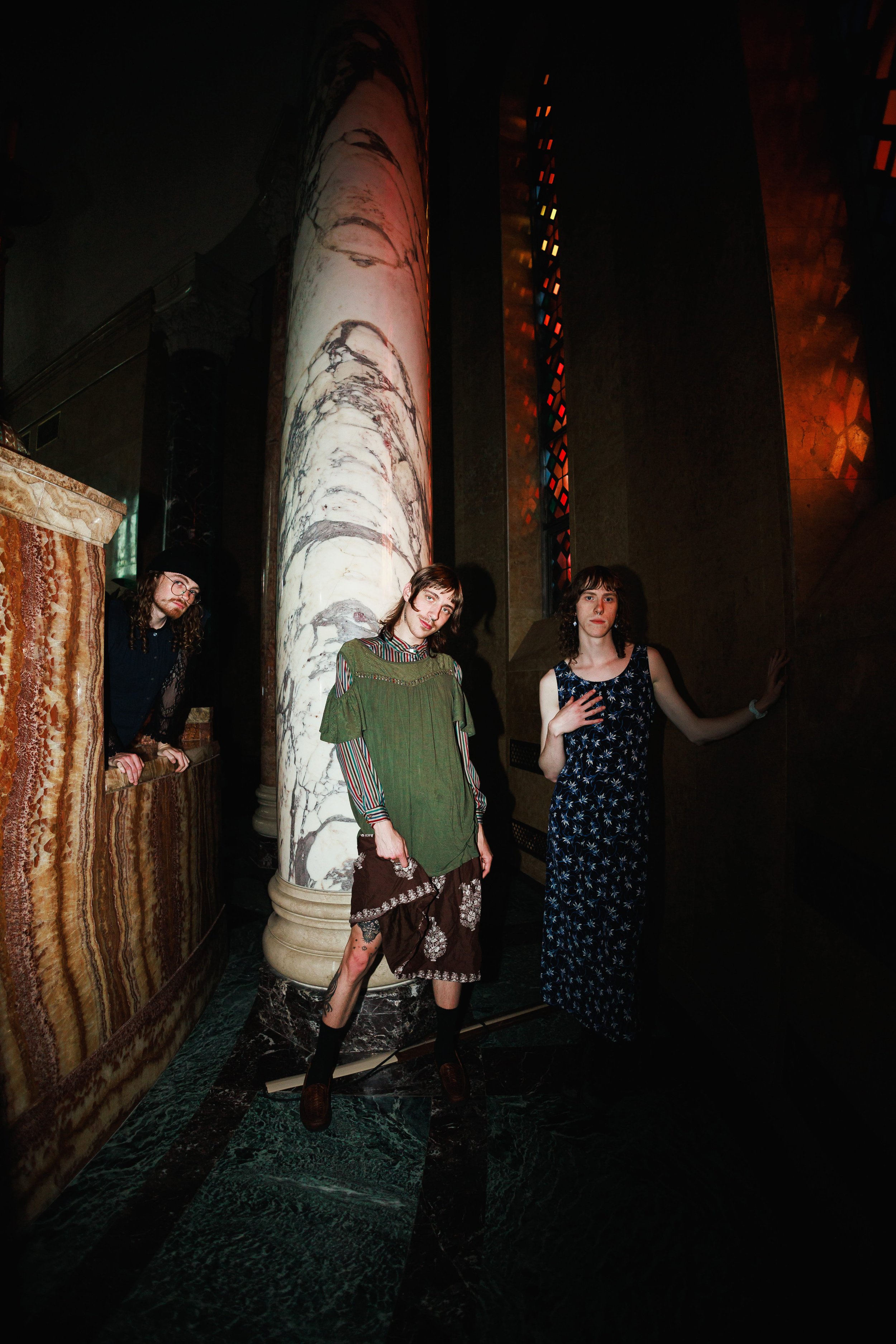 Four young people standing inside a dimly lit room with dark marble floors and large, decorative marble columns. One woman is wearing a long blue floral dress, another is in a green layered dress with striped sleeves, a third individual is wearing br