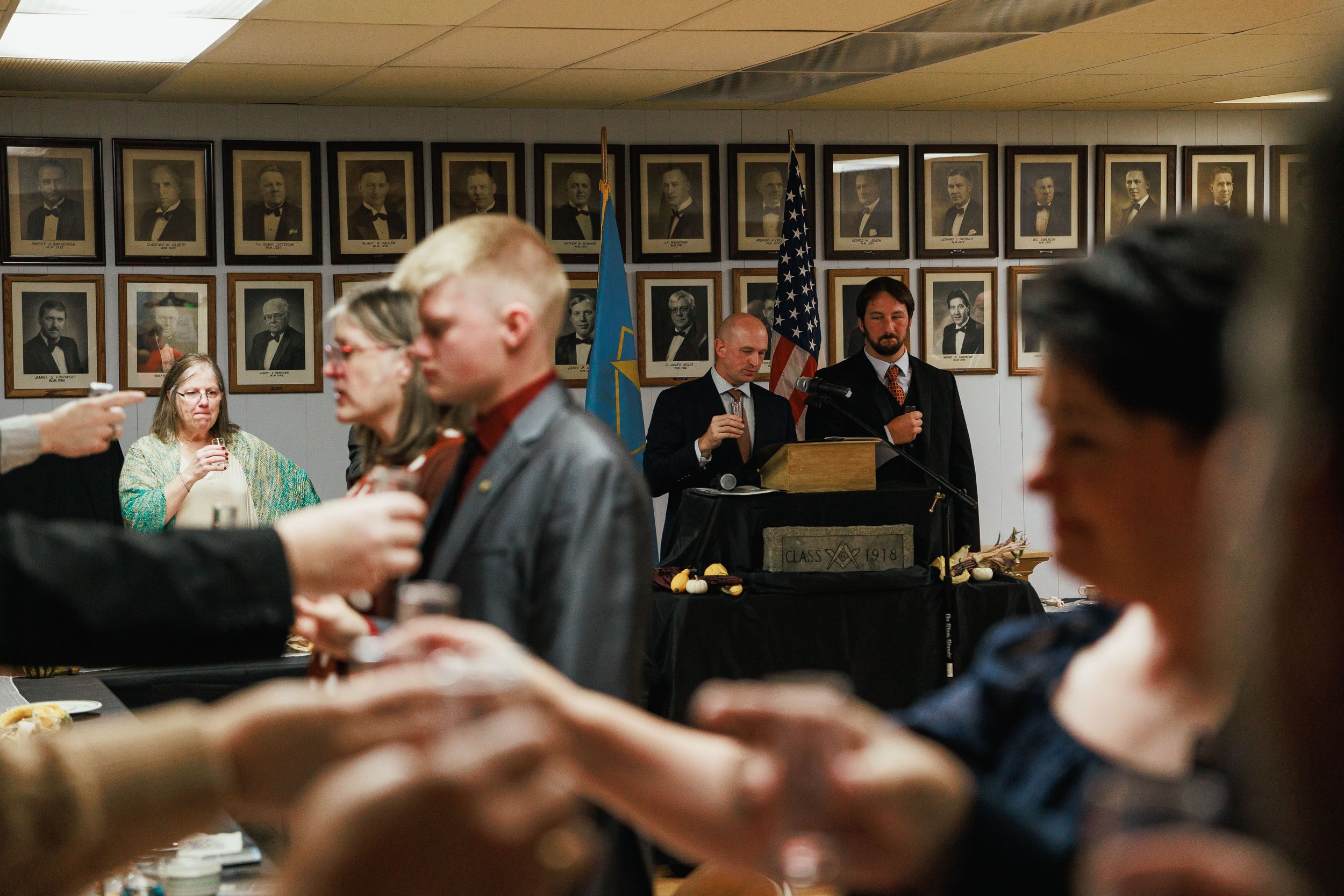 People attending a formal event in a hall decorated with framed portraits on the wall, with an American flag and a blue flag standing behind a podium at the front, where two men are speaking or presenting.