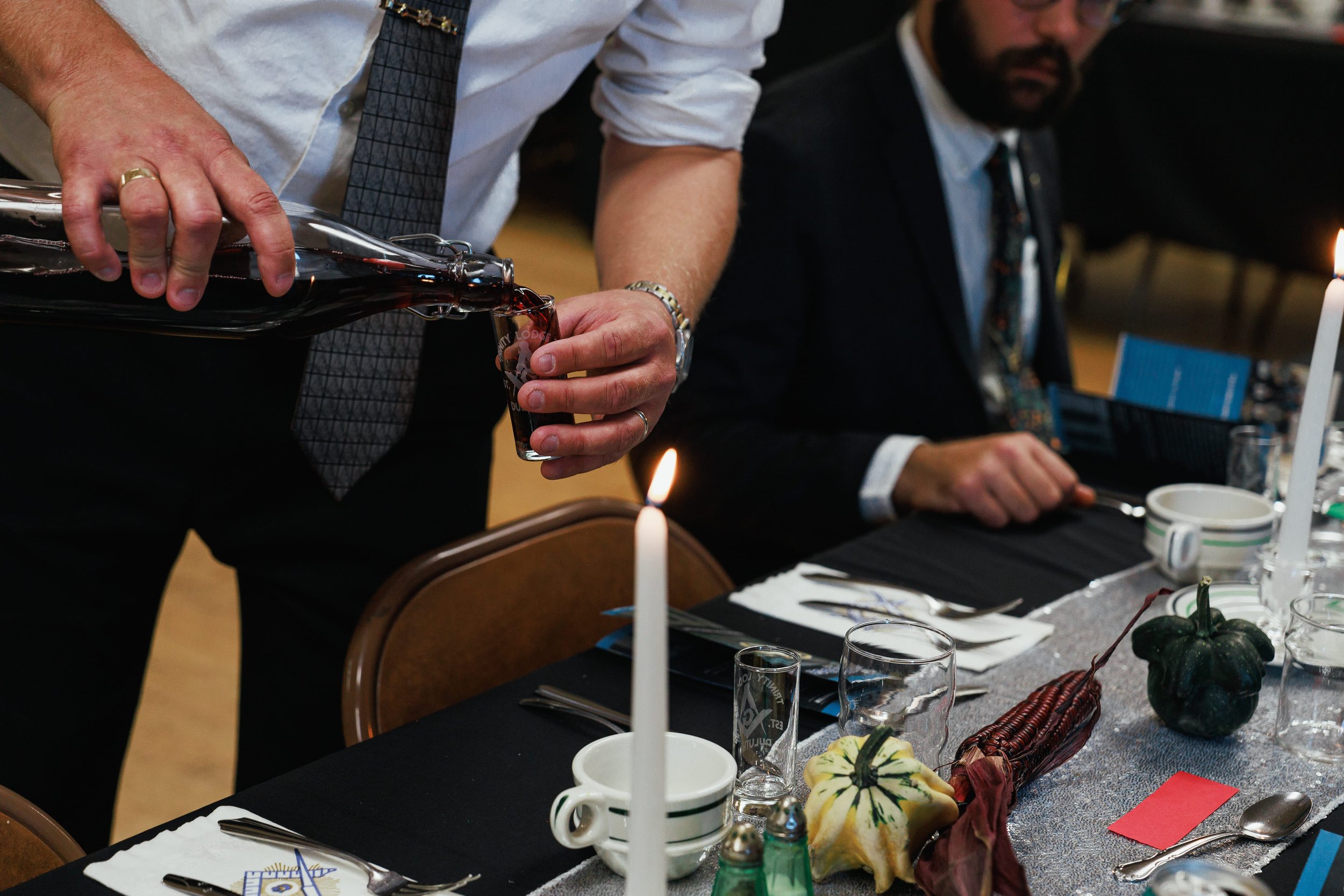 Man pouring red wine into a glass at a formal dinner table decorated with small pumpkins, candles, and tableware.