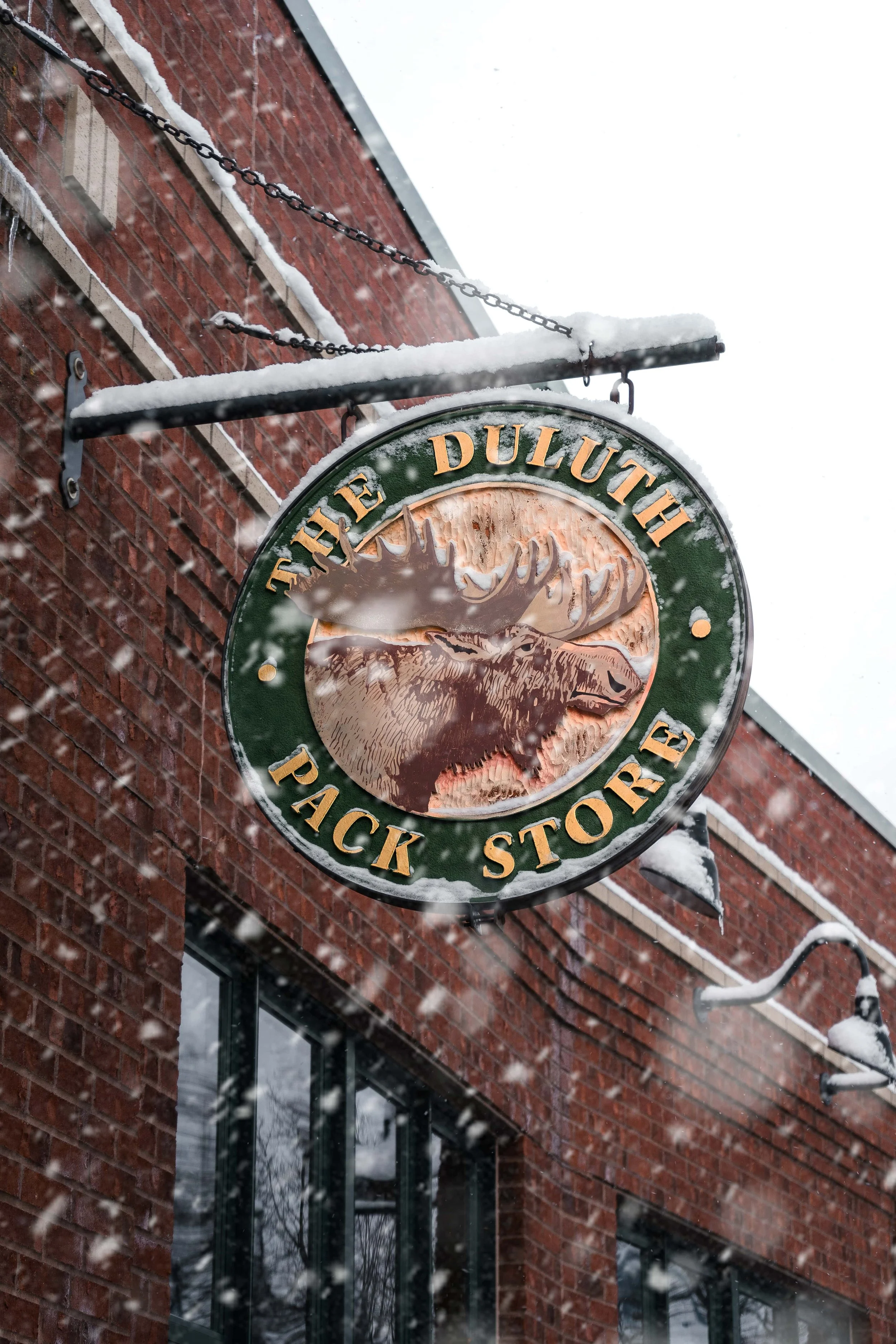 A circular sign hanging outside a brick building with snow on it. The sign says "The Duluth Pack Store" and features an image of a moose head.
