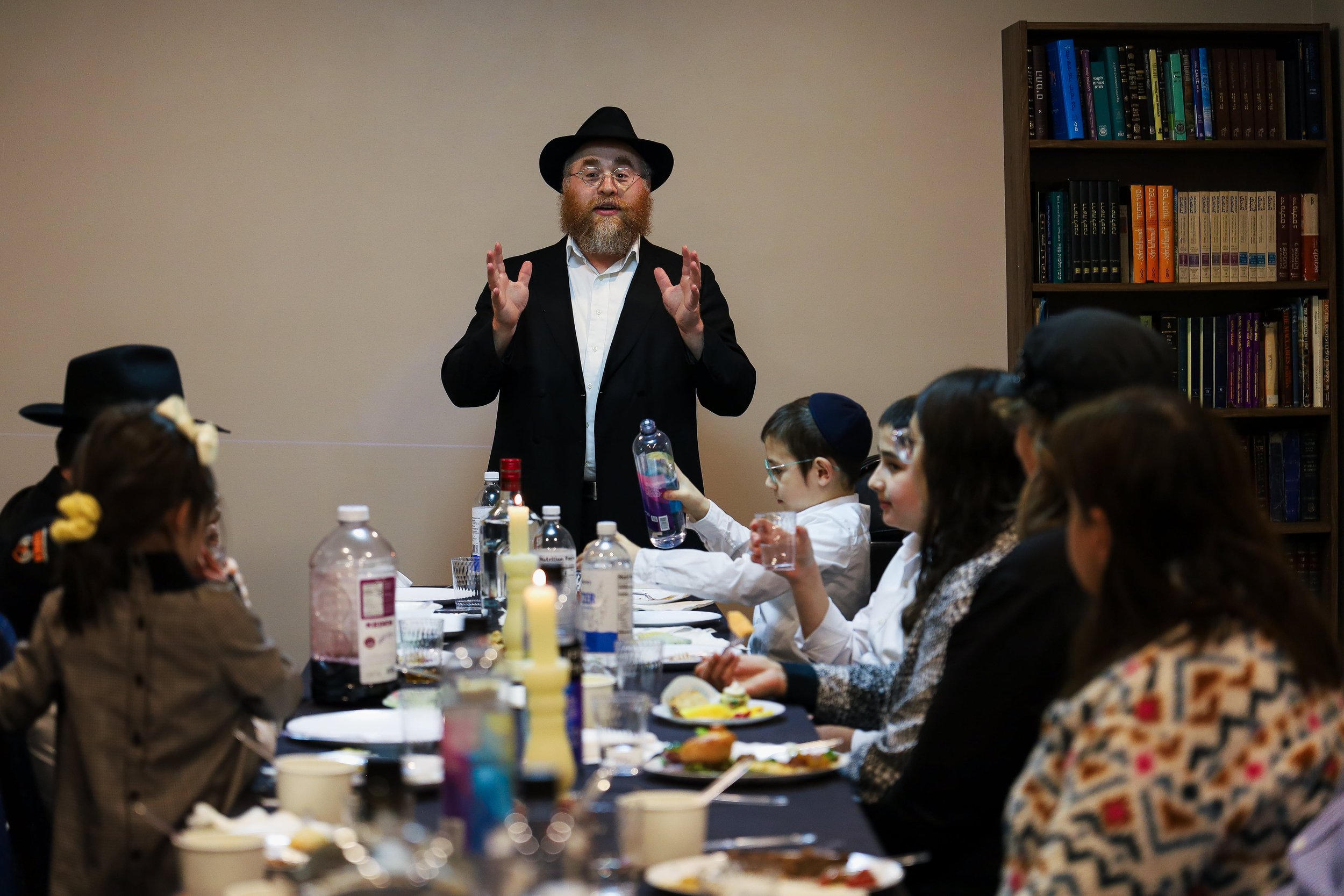 Man with beard, glasses, black hat, and black coat speaking to a group of children seated at a long table, with bookshelves in the background, during a dinner or event.