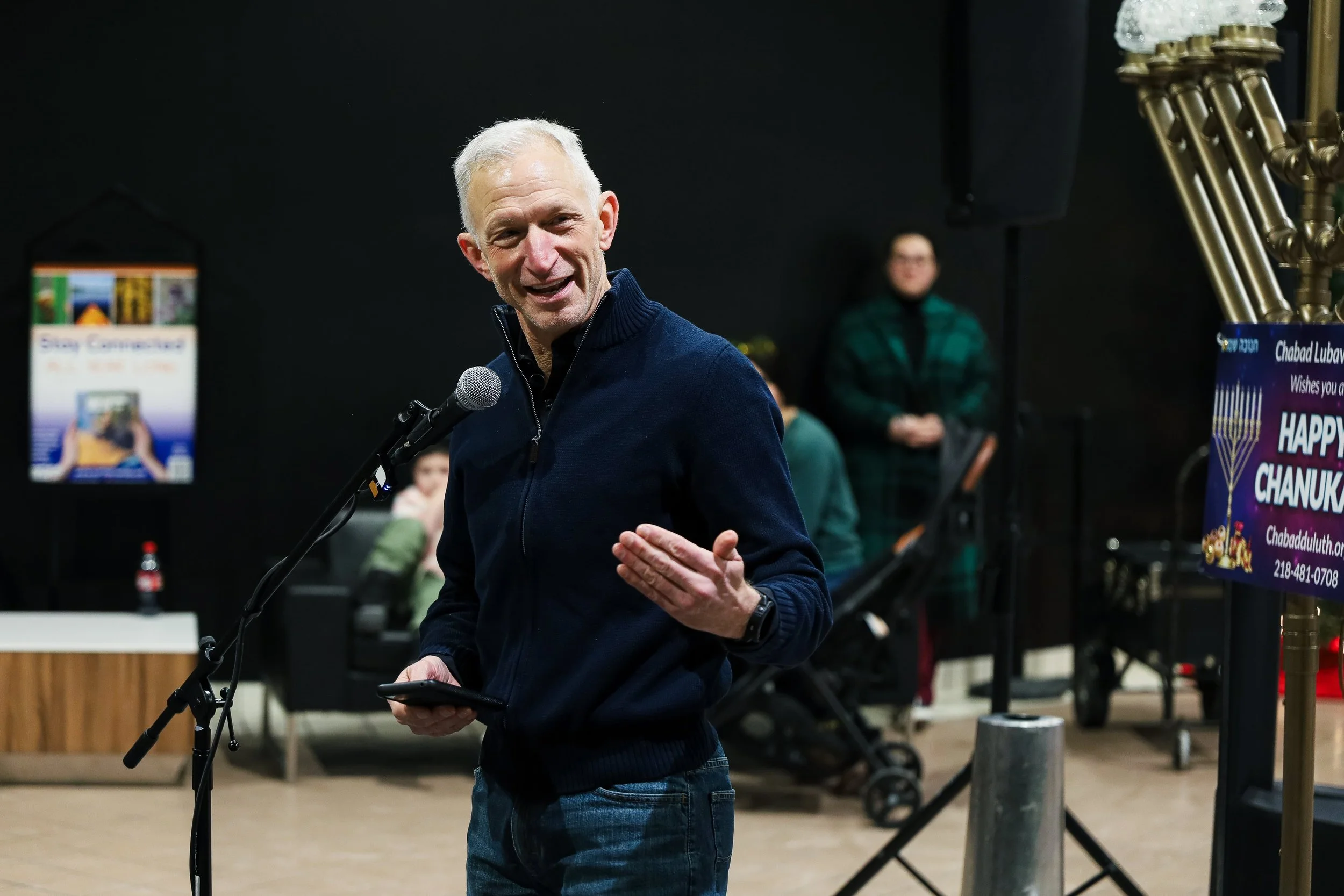 A man with short gray hair speaking into a microphone, holding a phone, at a Hanukkah celebration event. In the background, a woman and children watch, with Hanukkah decorations and a sign that reads 'Happy Chanukah'.