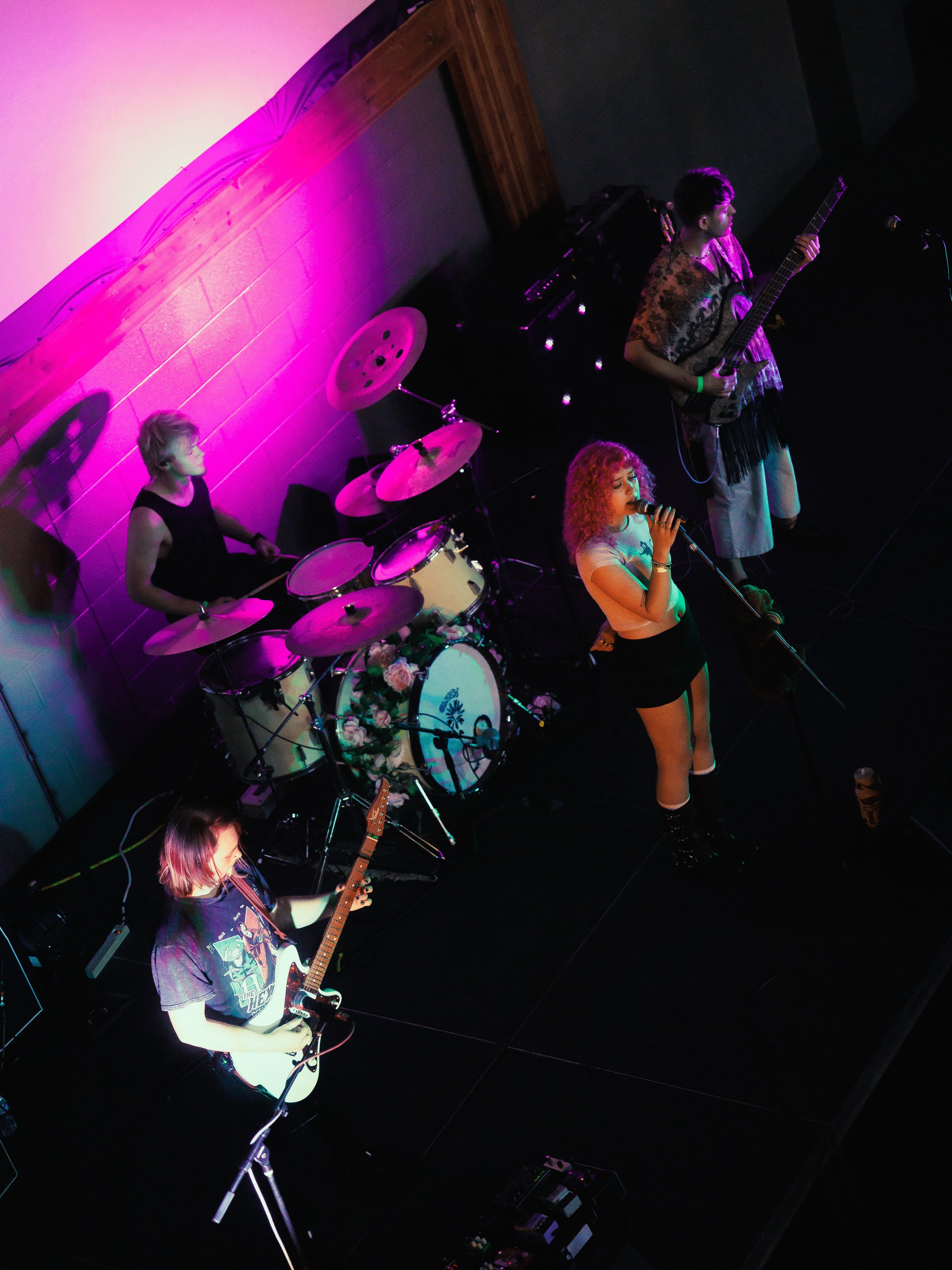 A band performing on stage with a female singer, a guitarist, a drummer, and a bassist, under colored stage lighting.