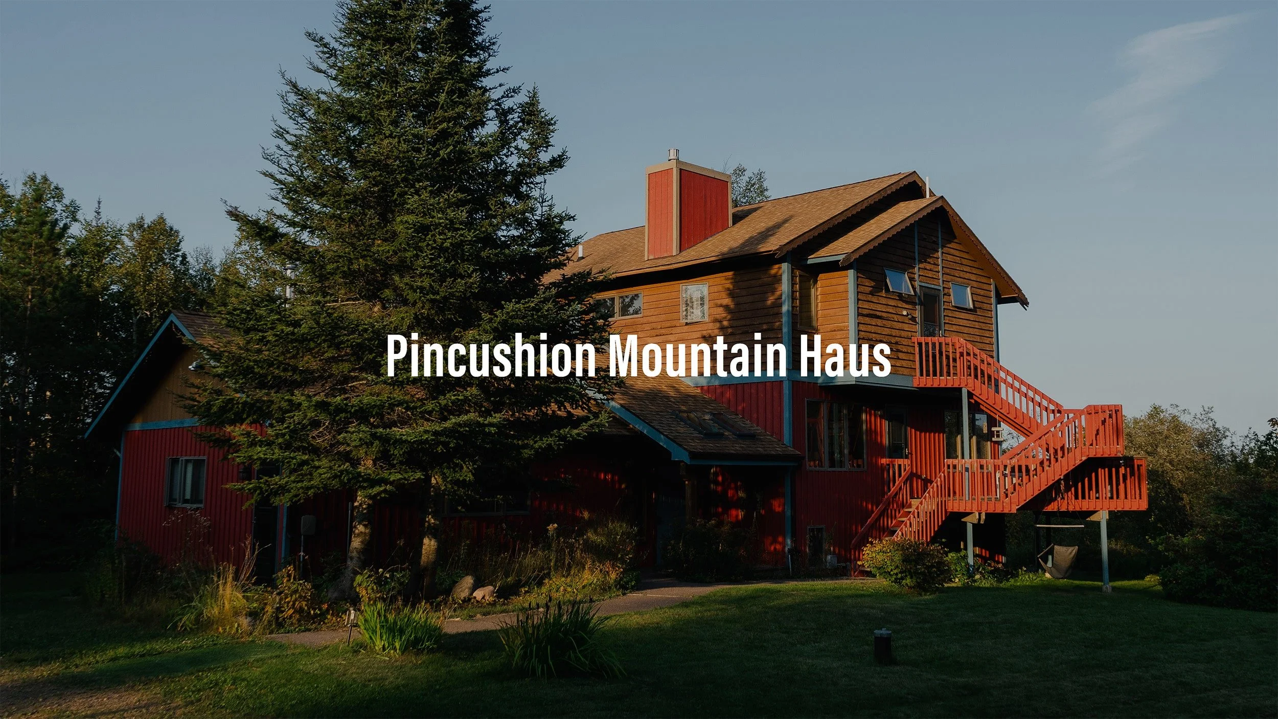 A multi-story mountain house painted red and brown, with a large tree nearby, set in lush green landscape under a clear blue sky.