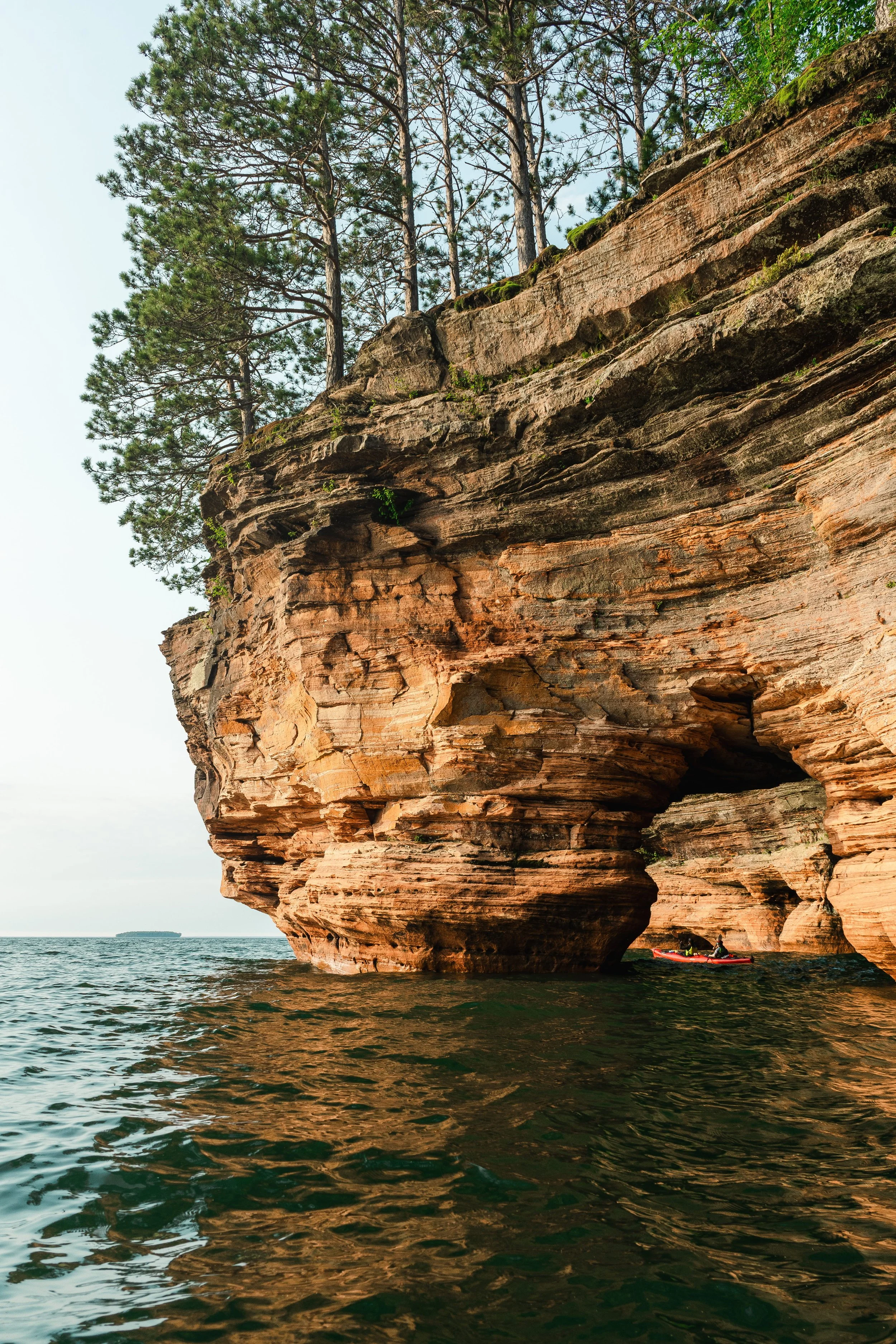 A large reddish-brown rocky cliff face rising from the water with pine trees on top. A person is kayaking near the base of the cliff, and a distant ship is visible on the horizon.