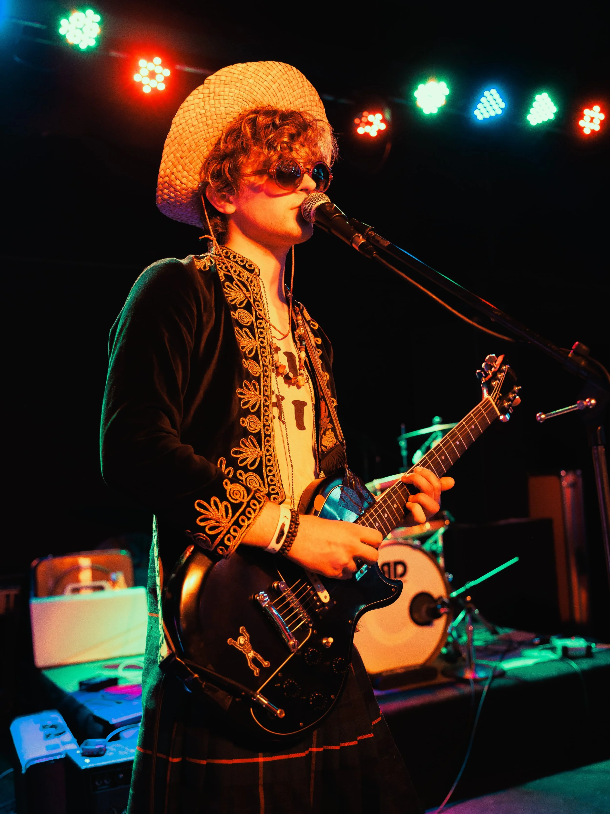 A person performing on stage with a guitar, wearing a wide-brimmed straw hat, sunglasses, and a jacket with decorative embroidery, under stage lights.