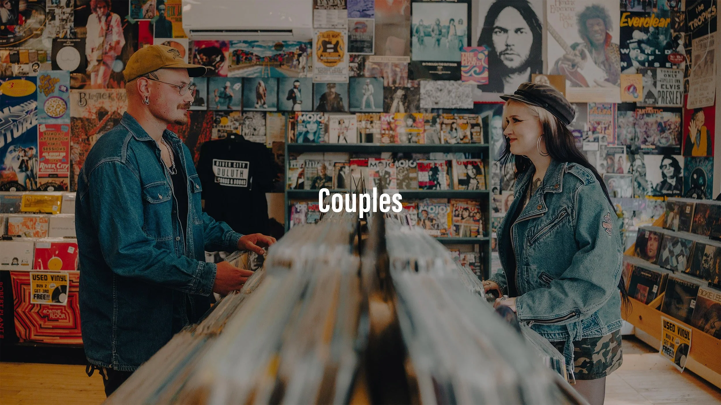 A couple browsing records in a record store, with posters and album covers on the walls, and shelves filled with vinyl and music magazines.