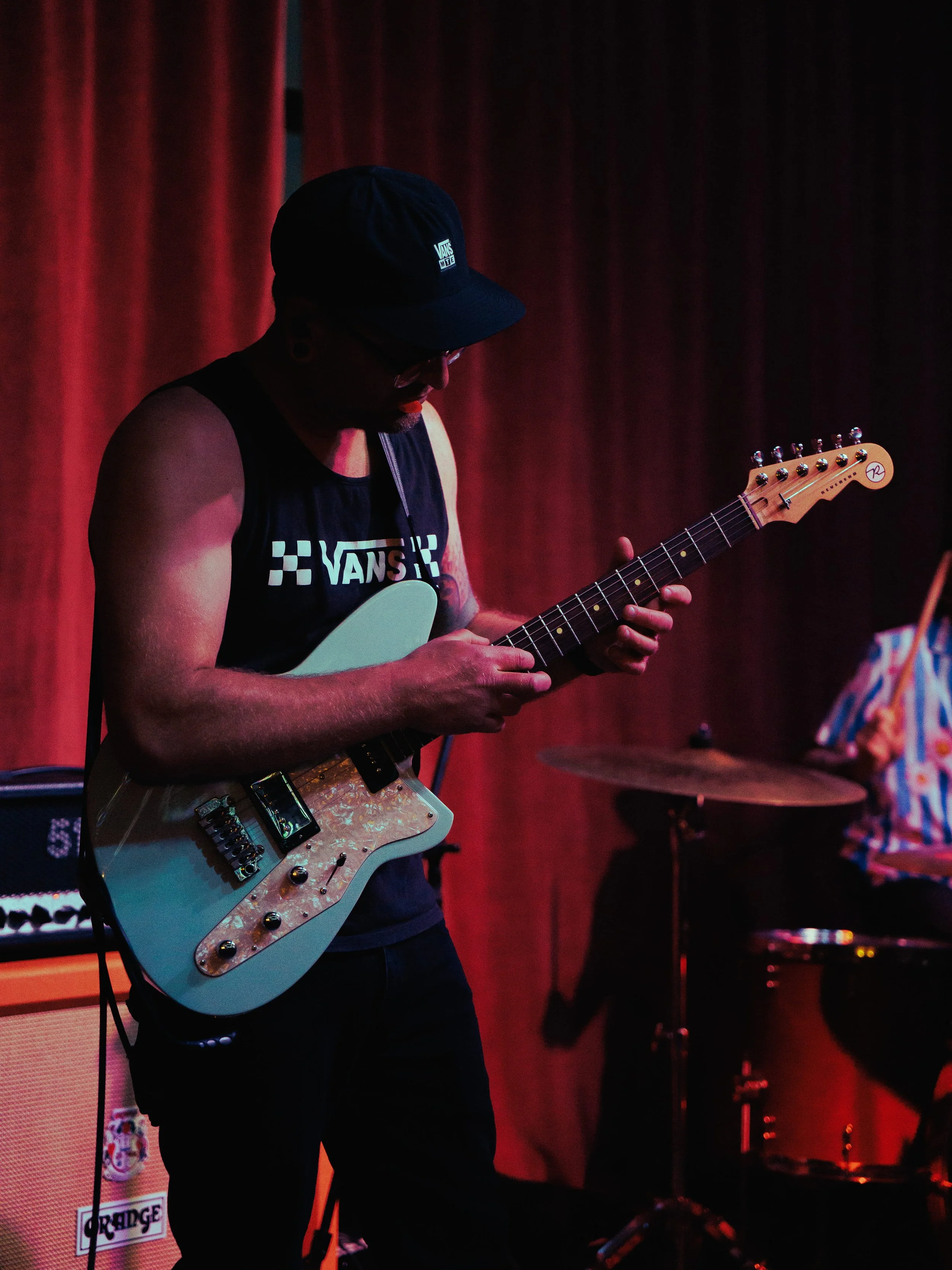 A person playing an electric guitar on stage, wearing a black Vans cap and sleeveless shirt, with red curtain background and drum set nearby.
