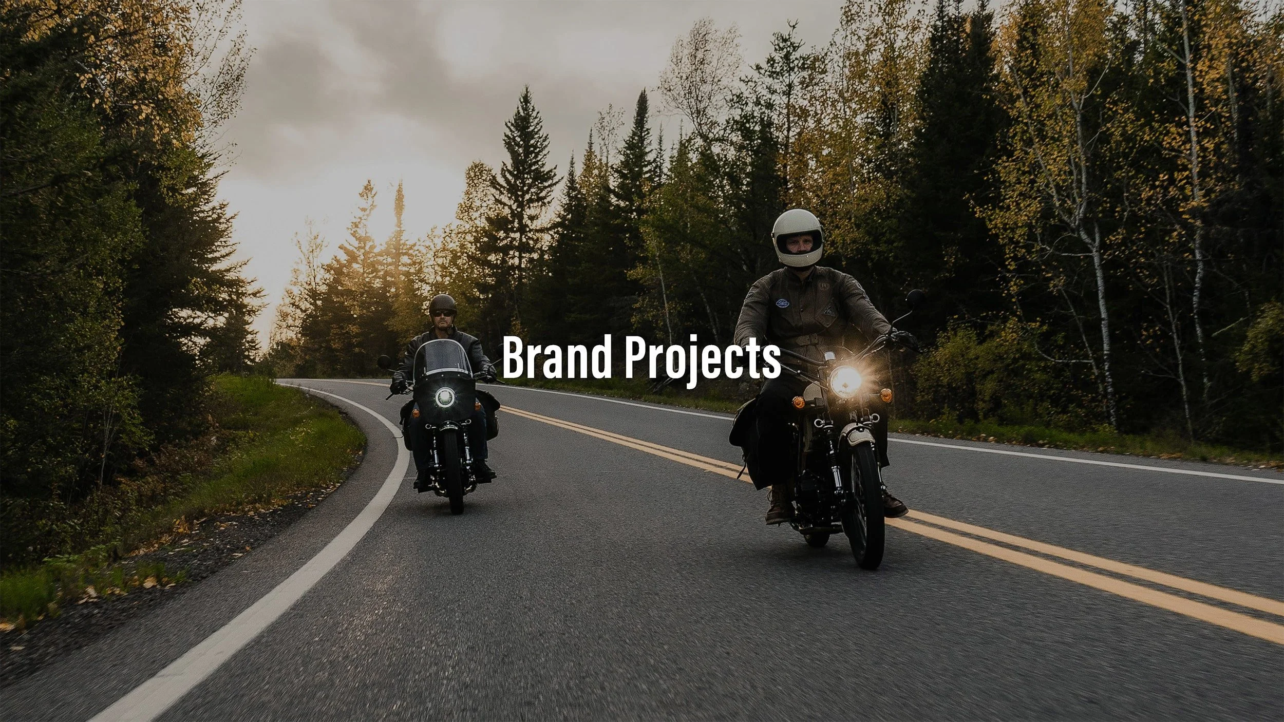 Two motorcyclists riding on a winding road through a forested area with trees and bushes, with the sun setting or rising in the background. The text 'Brand Projects' is overlaid on the image.