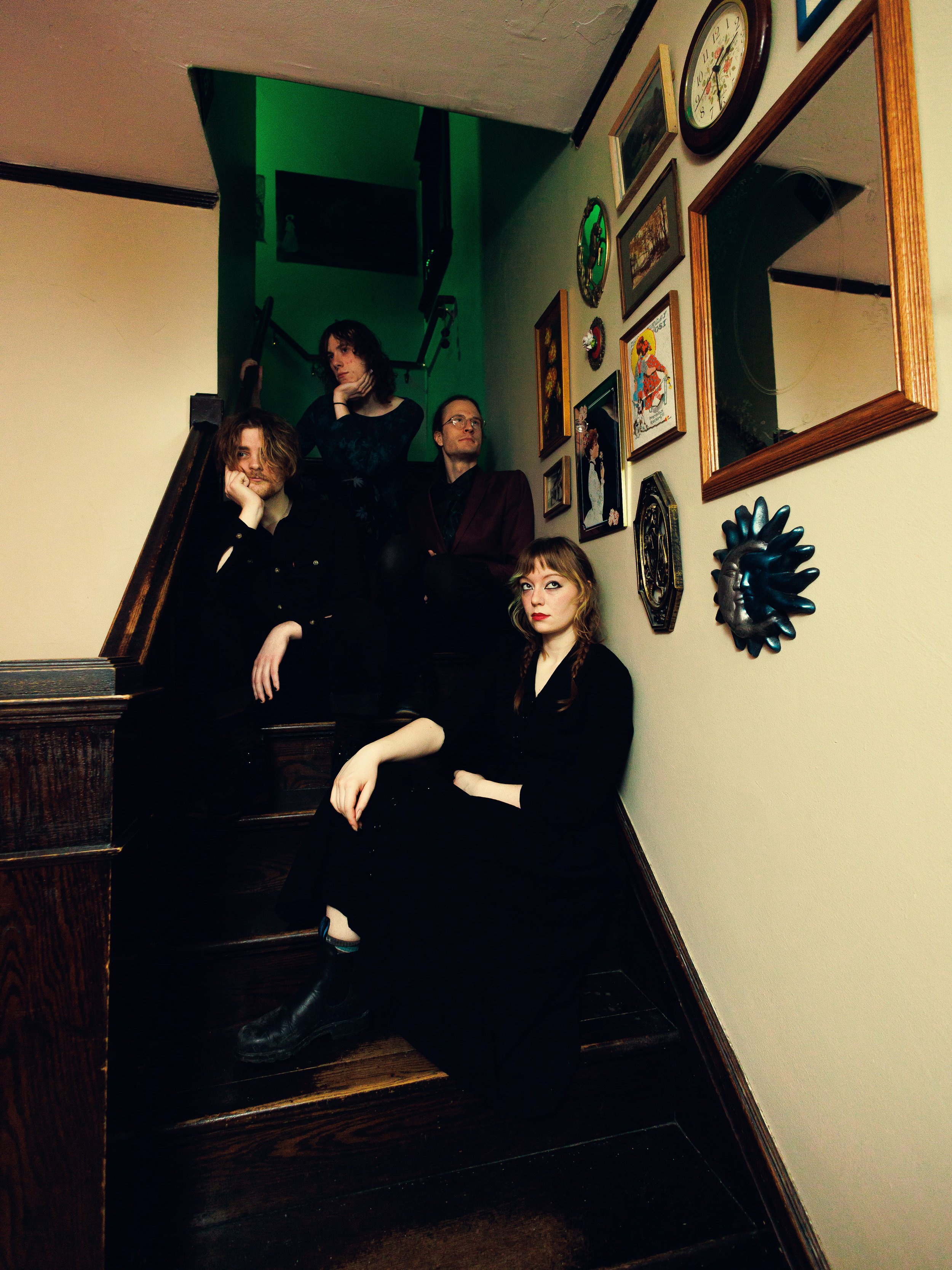 Four people are on a staircase in a house, with framed pictures and a clock on the wall. The person at the top appears to be a woman with dark hair, resting her chin on her hand. The person second from the bottom is a man with glasses, crossing his a