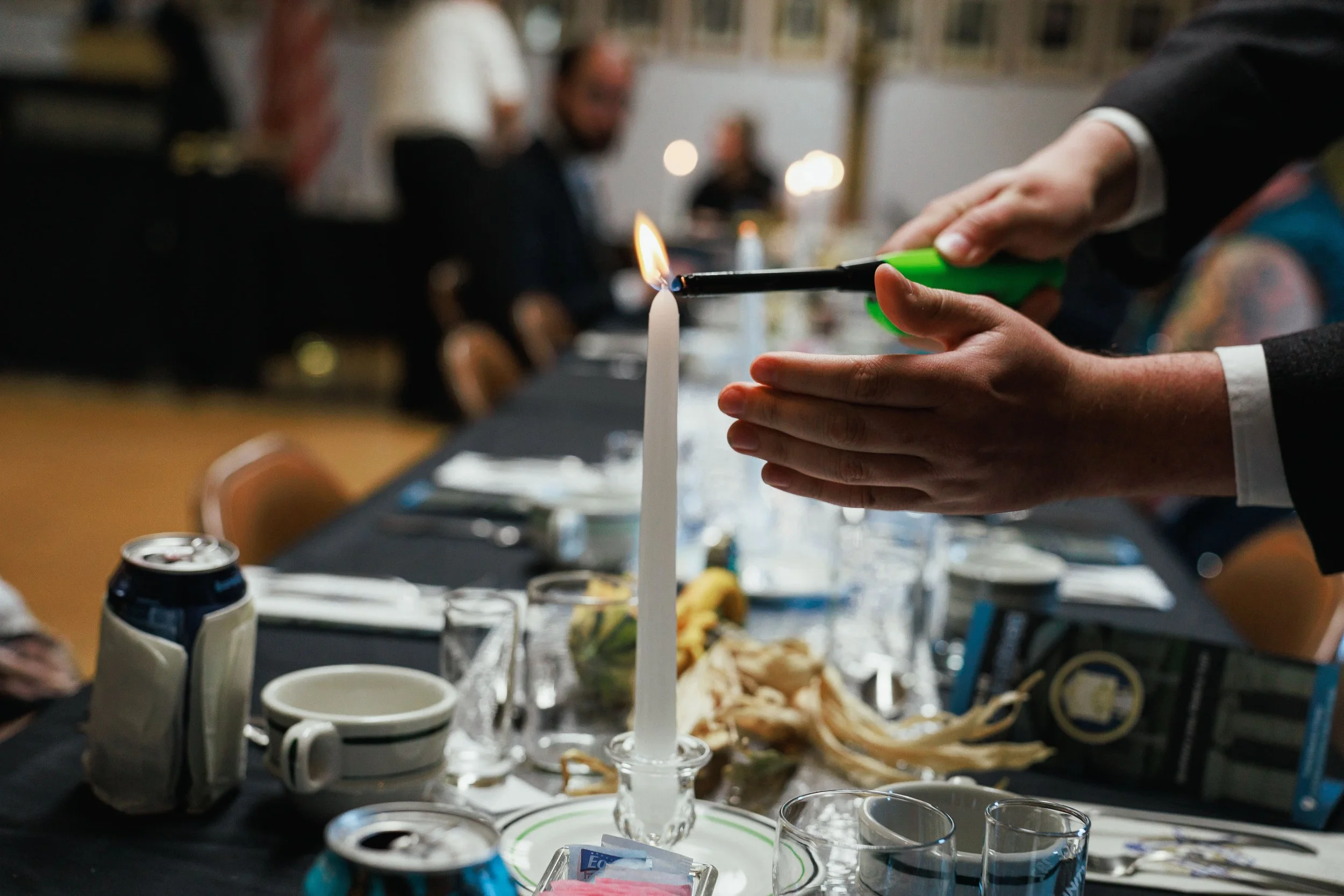 Person lighting a large white candle on a table during a formal event, with others seated in the background.