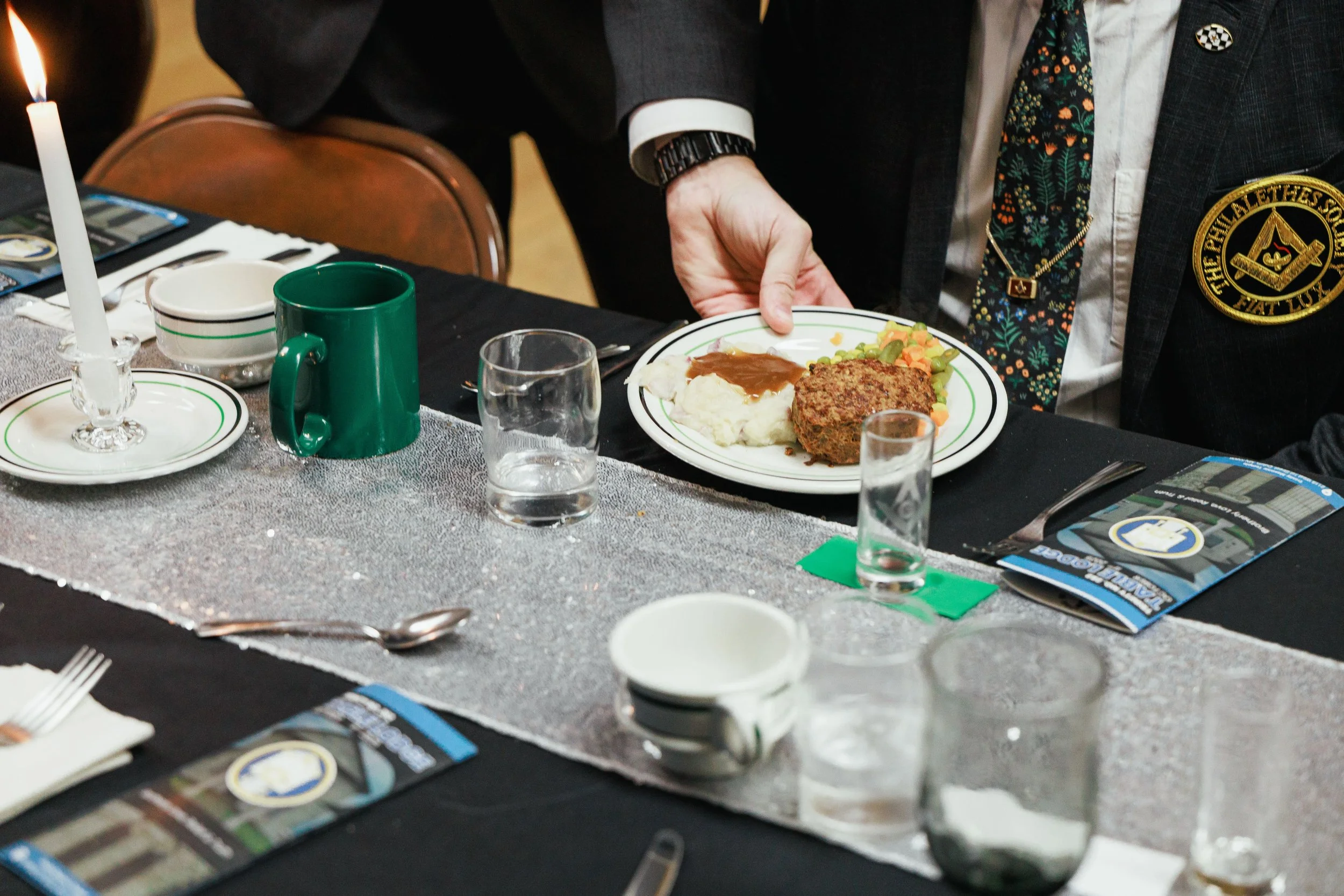 A man dressed in a suit and patterned tie serving food on a plate at a table set for a meal, with various dishes, glasses, cups, and silverware.