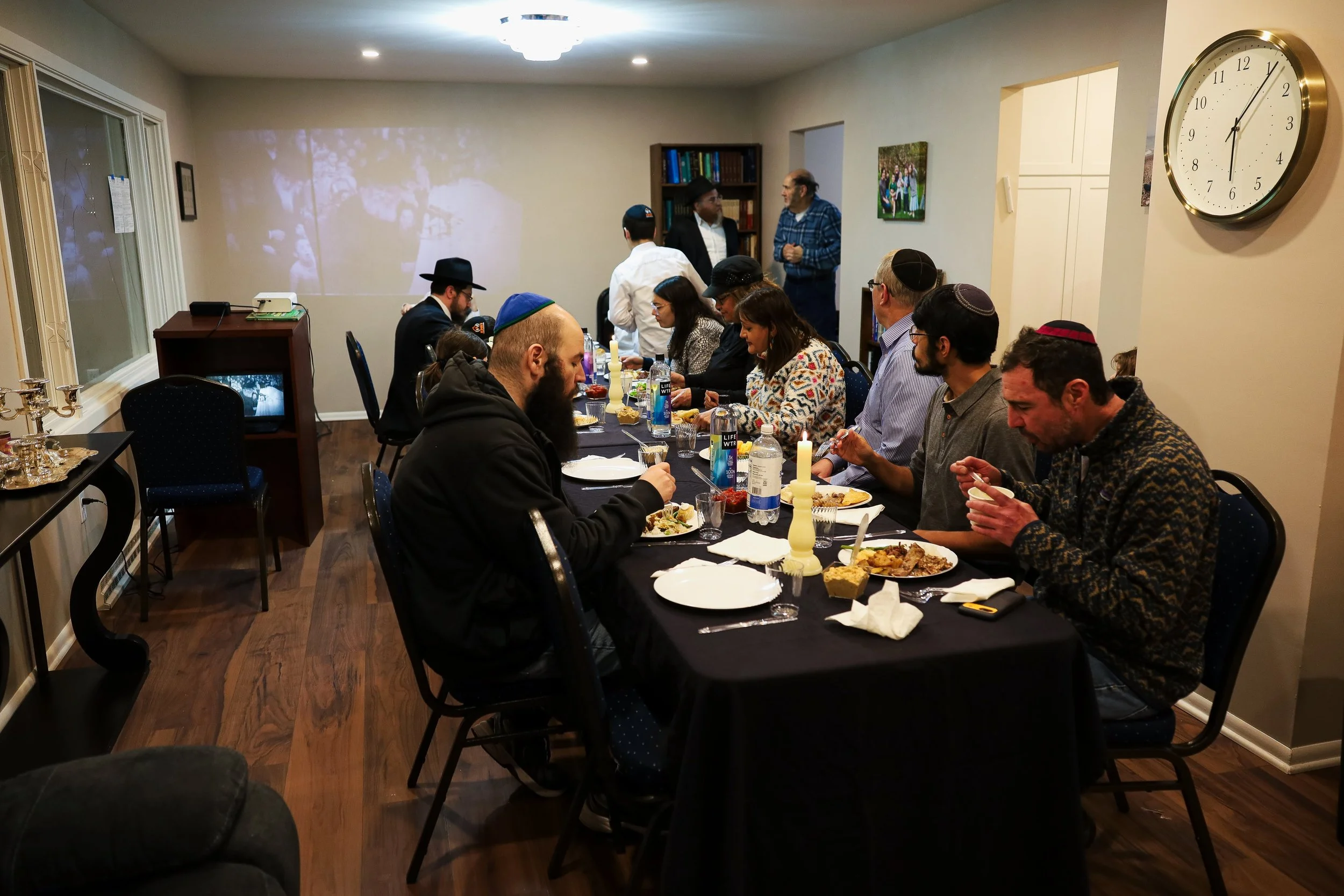 A group of people are gathered around a long dining table having a meal in a private room. Some are wearing kippahs, indicating Jewish tradition. The room is decorated simply, with a large wall clock and bookshelves in the background. Several people 