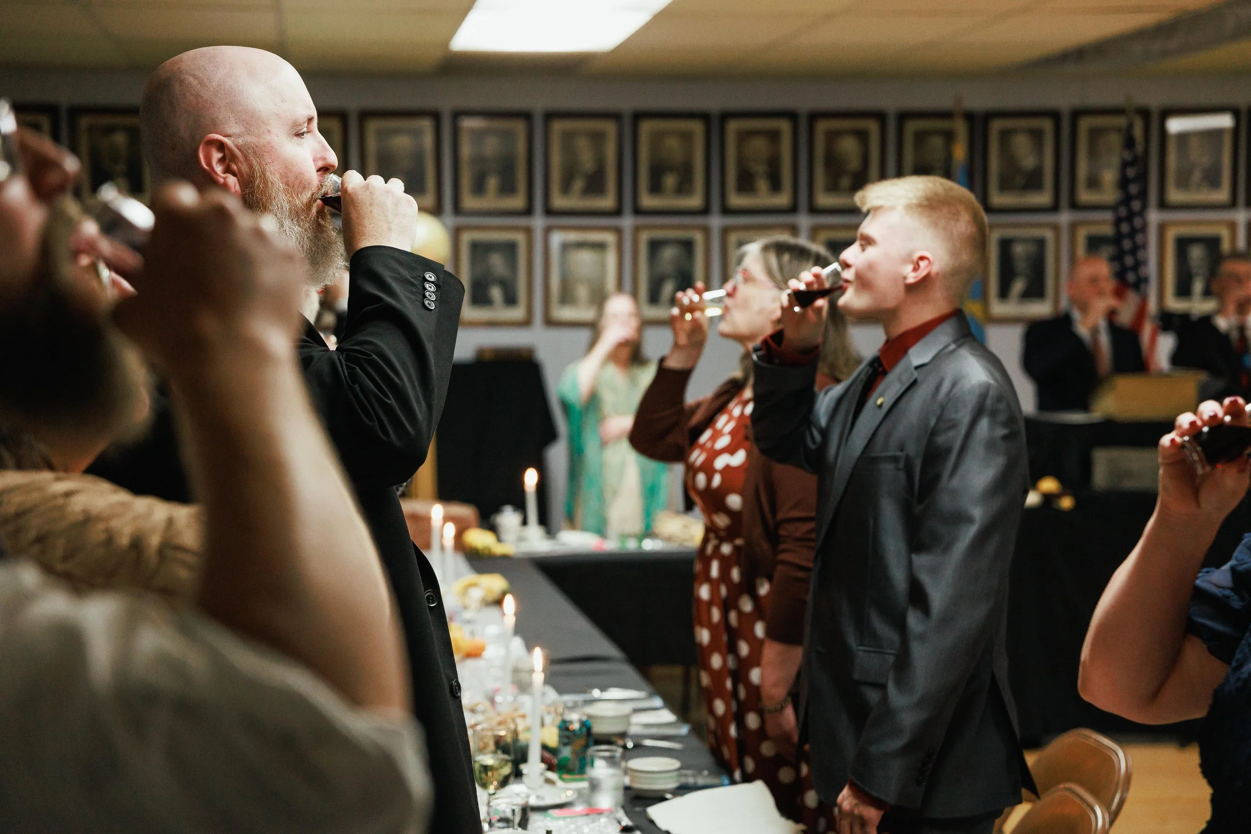 People at a formal gathering toasting with drinks in a decorated room with framed photographs on the wall.