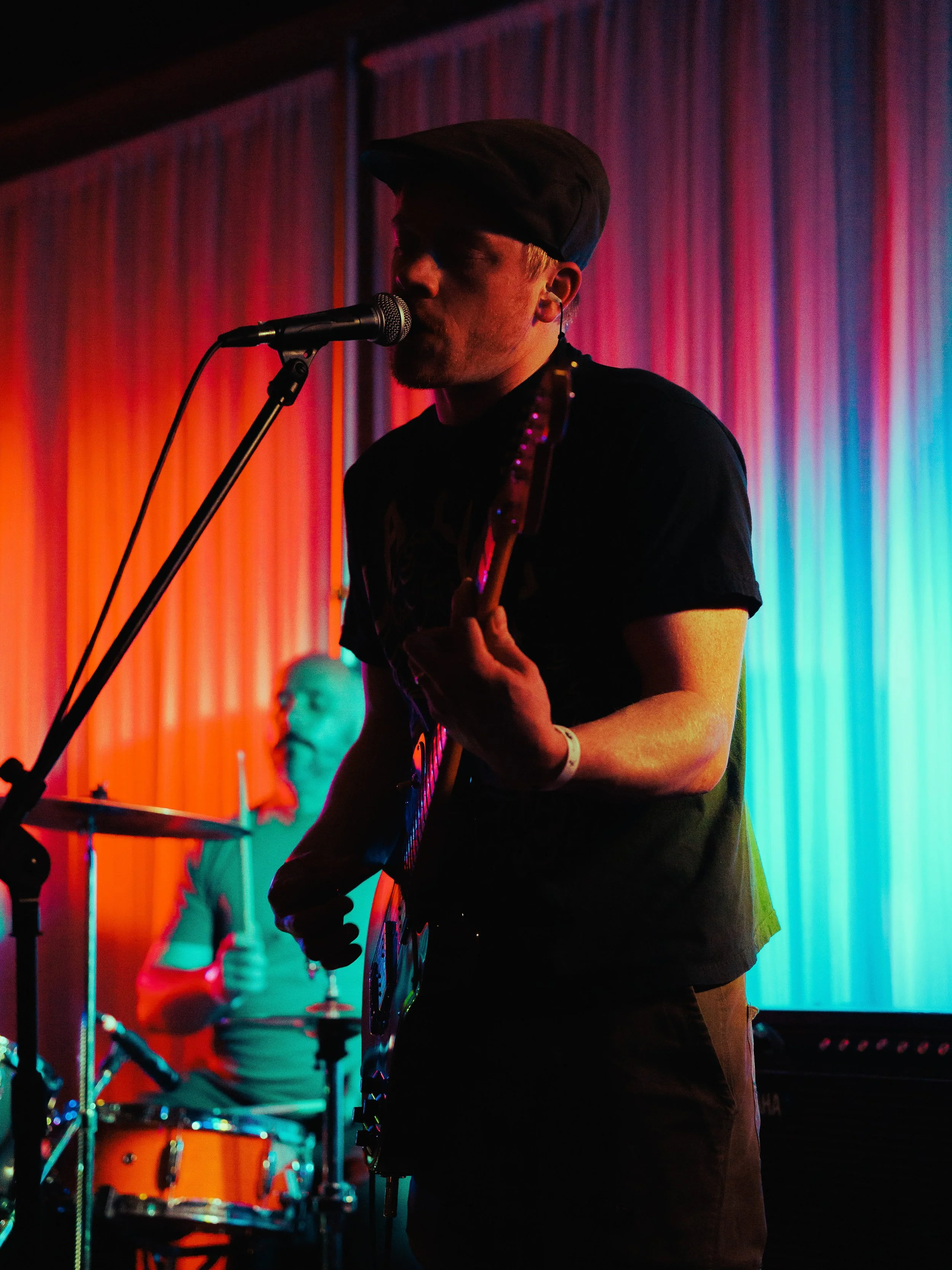 A musician performing on stage with colorful lighting, playing an electric guitar, with a drum set in the background and a person with glasses sitting behind him.