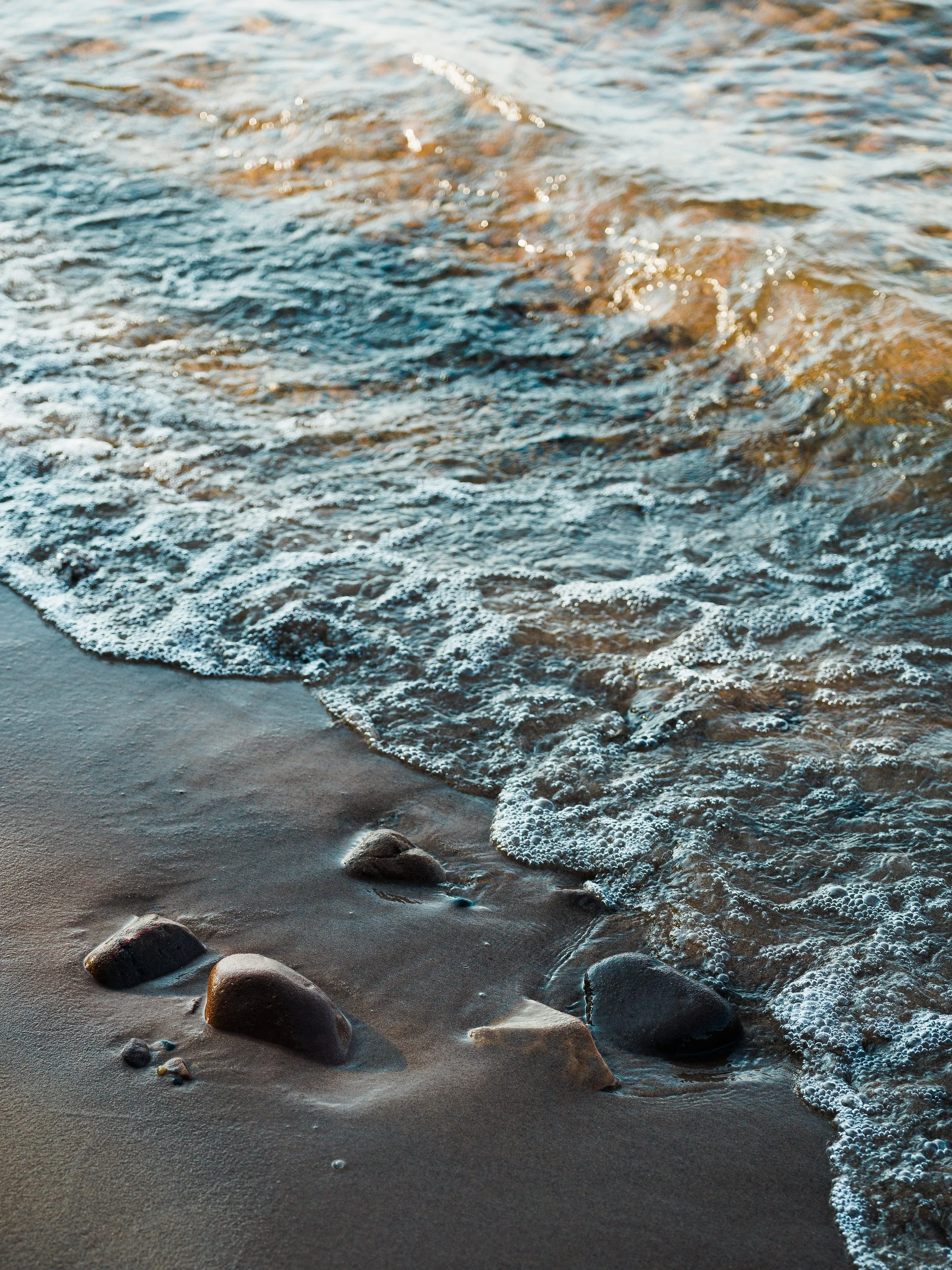 Waves lapping rocks on a sandy beach at sunset.
