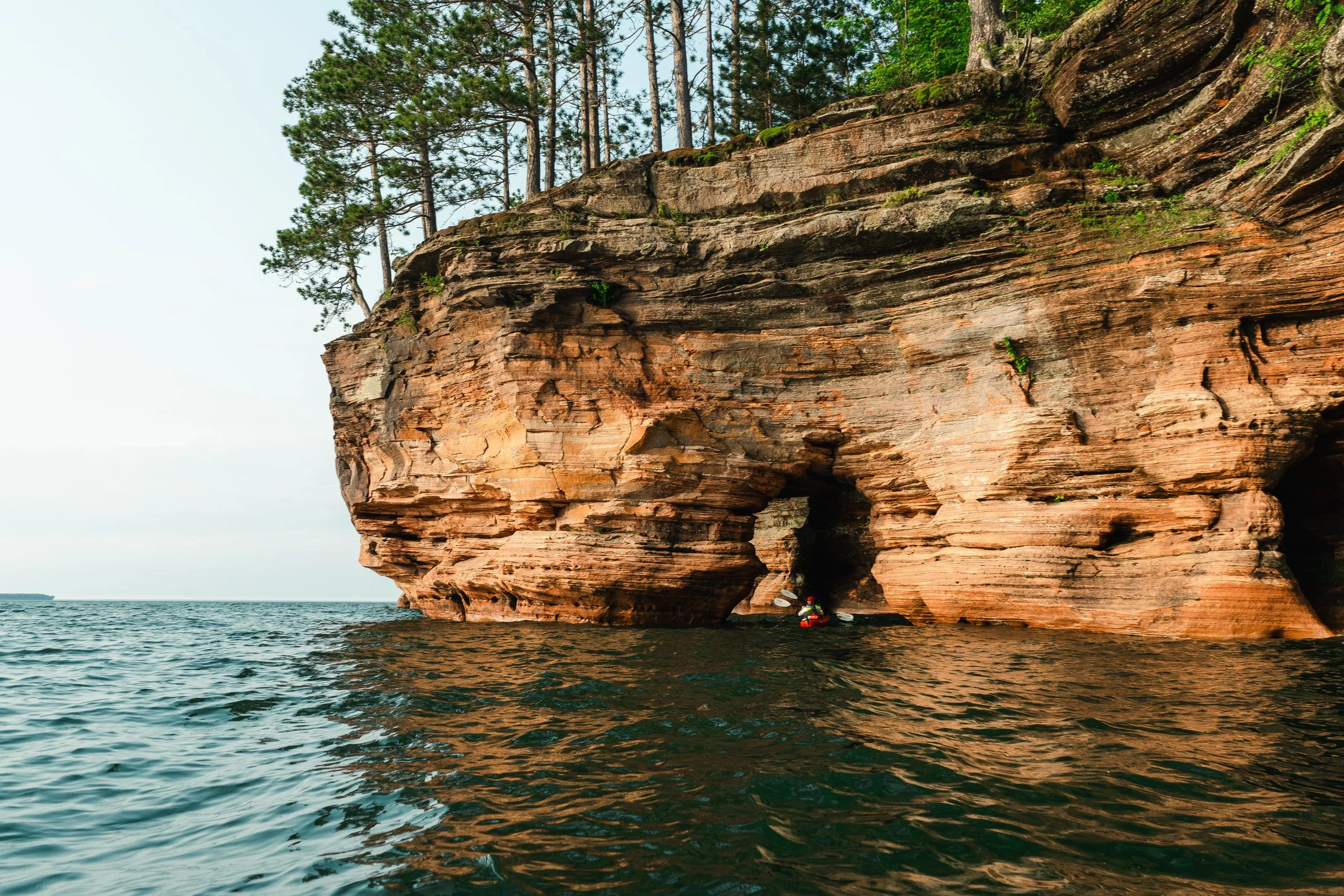 A large rock formation with trees growing on top, extending into the water with a small waterfall. Two people are kayaking near the base of the rock.
