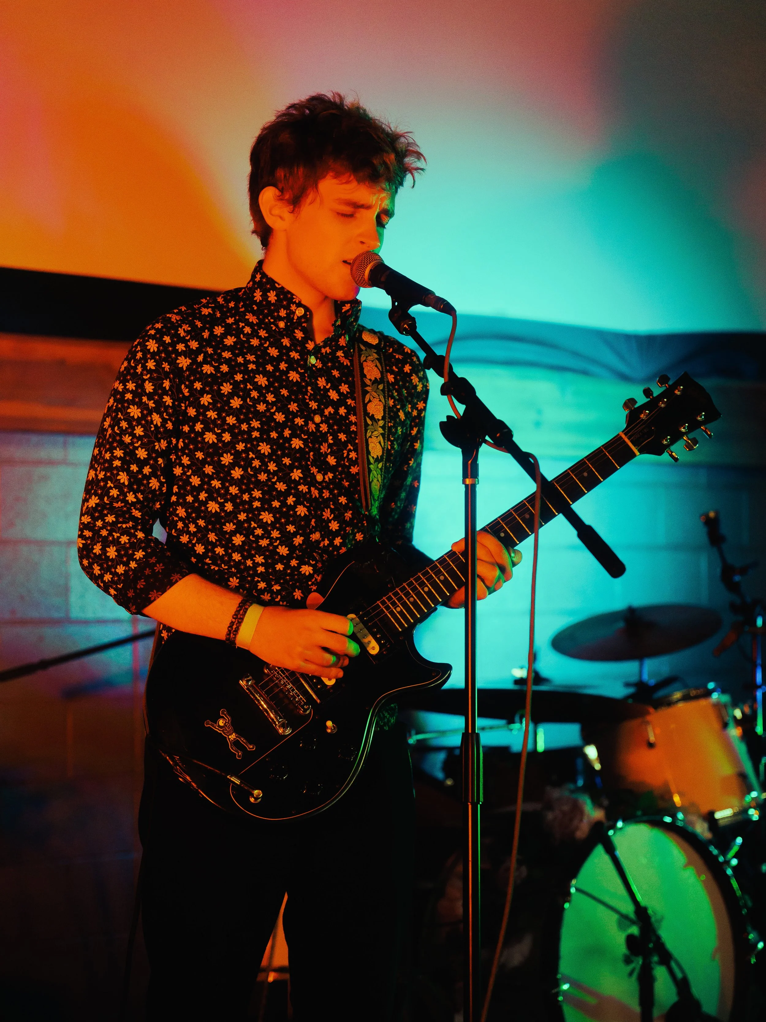 A young male musician is performing on stage, singing into a microphone and playing an electric guitar with a black body and roses decoration. He has short, wavy hair and is wearing a black shirt pattern with small orange flowers. The stage has color