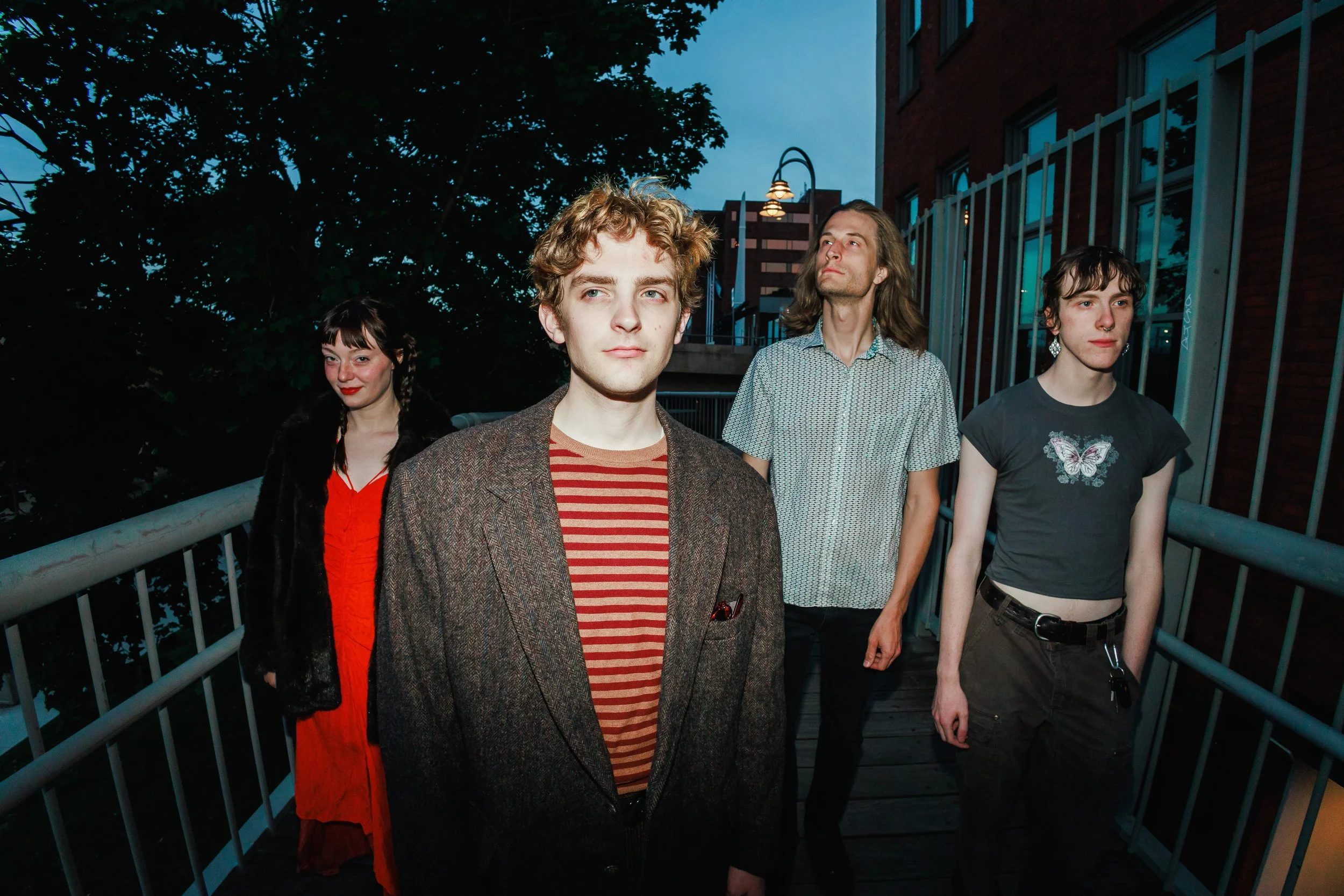 Four young adults standing outdoors on a balcony or rooftop at dusk, with city buildings and trees in the background. They are dressed casually, with one woman wearing a red dress, a man in the foreground wearing a brown blazer and striped shirt, ano
