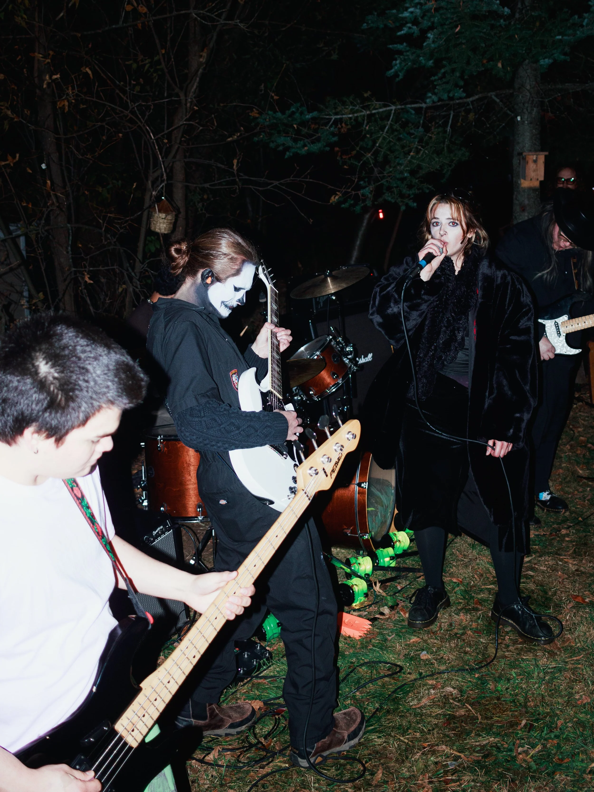 A band performs outdoors at night, featuring a woman singing with a microphone, musicians playing guitars, and a drummer in the background, surrounded by trees.