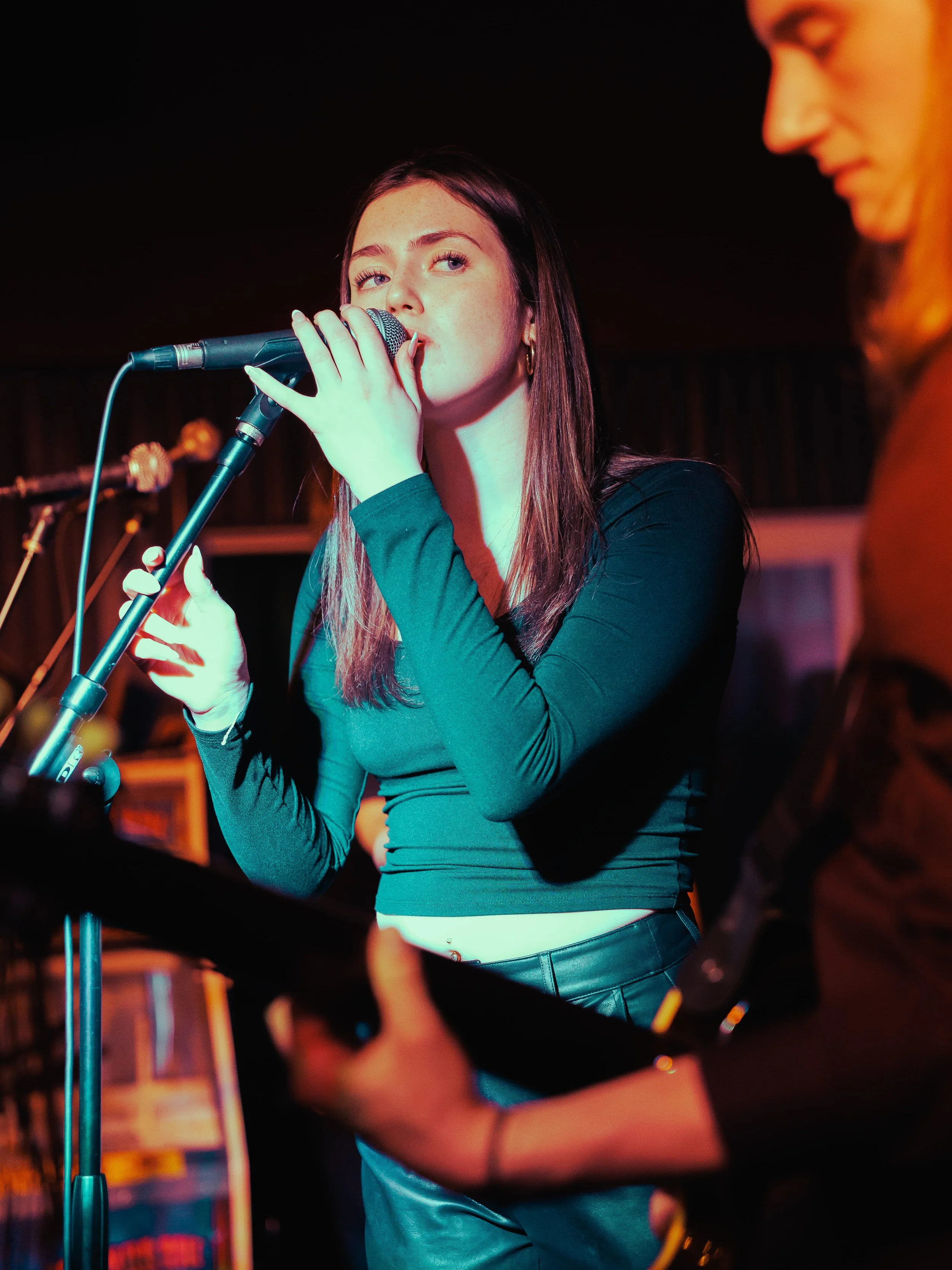 A young woman with long brown hair and hoop earrings singing into a microphone on stage, with a person in the foreground playing a guitar