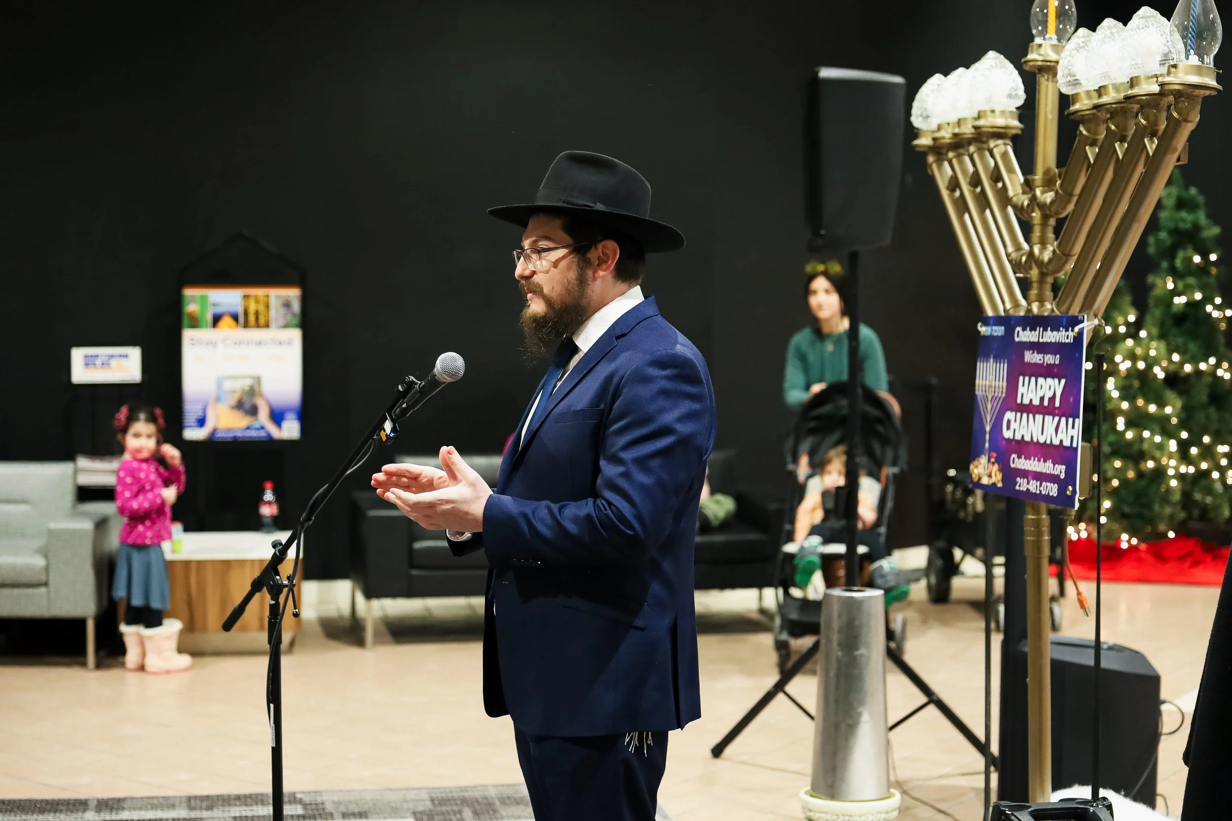 A man wearing a black hat, glasses, and a blue suit is speaking into a microphone at a Hanukkah celebration, with a menorah and a Christmas tree in the background.