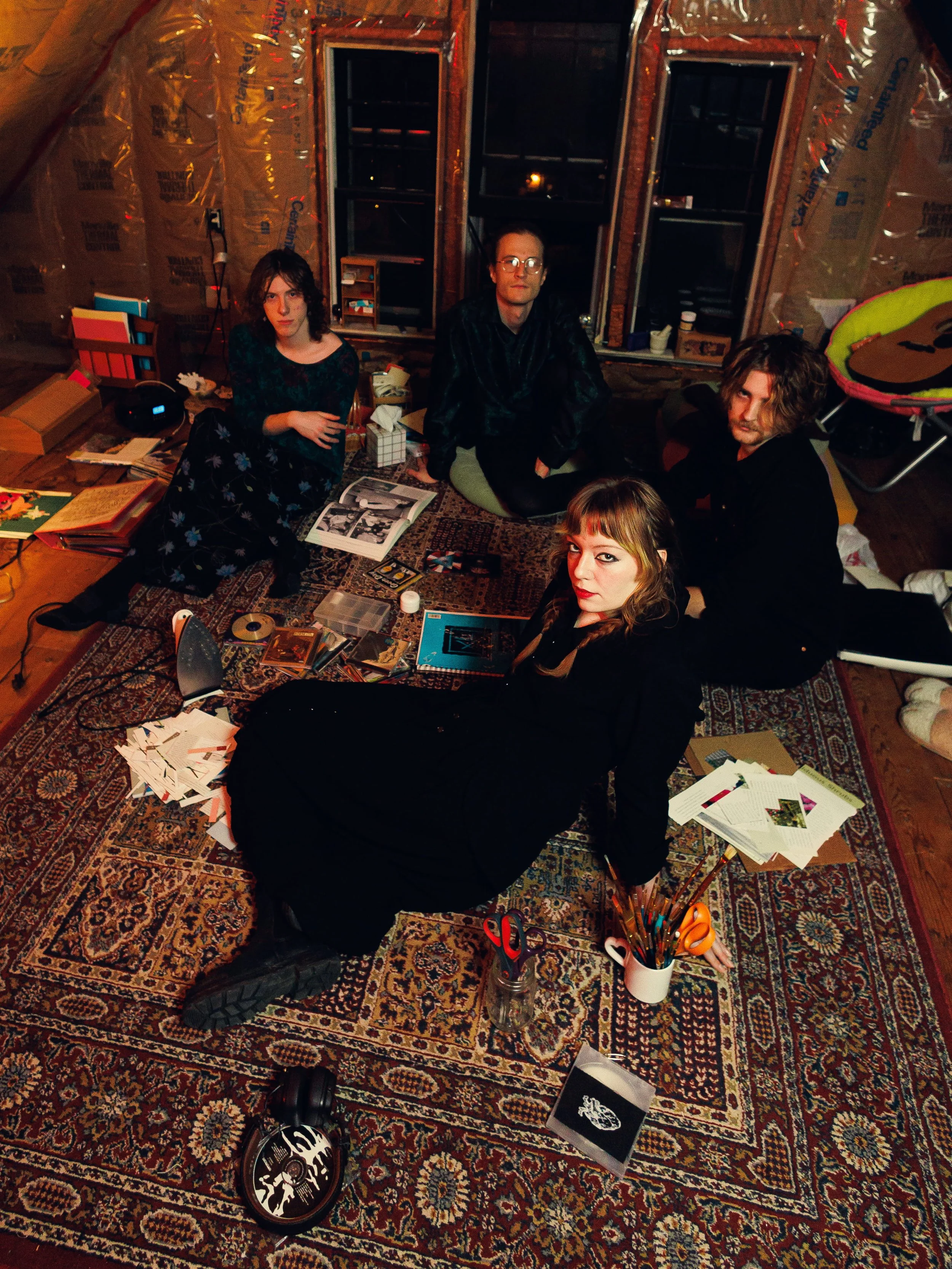 A group of four young adults in a cluttered, unfinished room with insulation on the walls, sitting and lying on a patterned carpet. They are surrounded by books, papers, art supplies, and various objects, including scissors, a skate helmet, and a gui