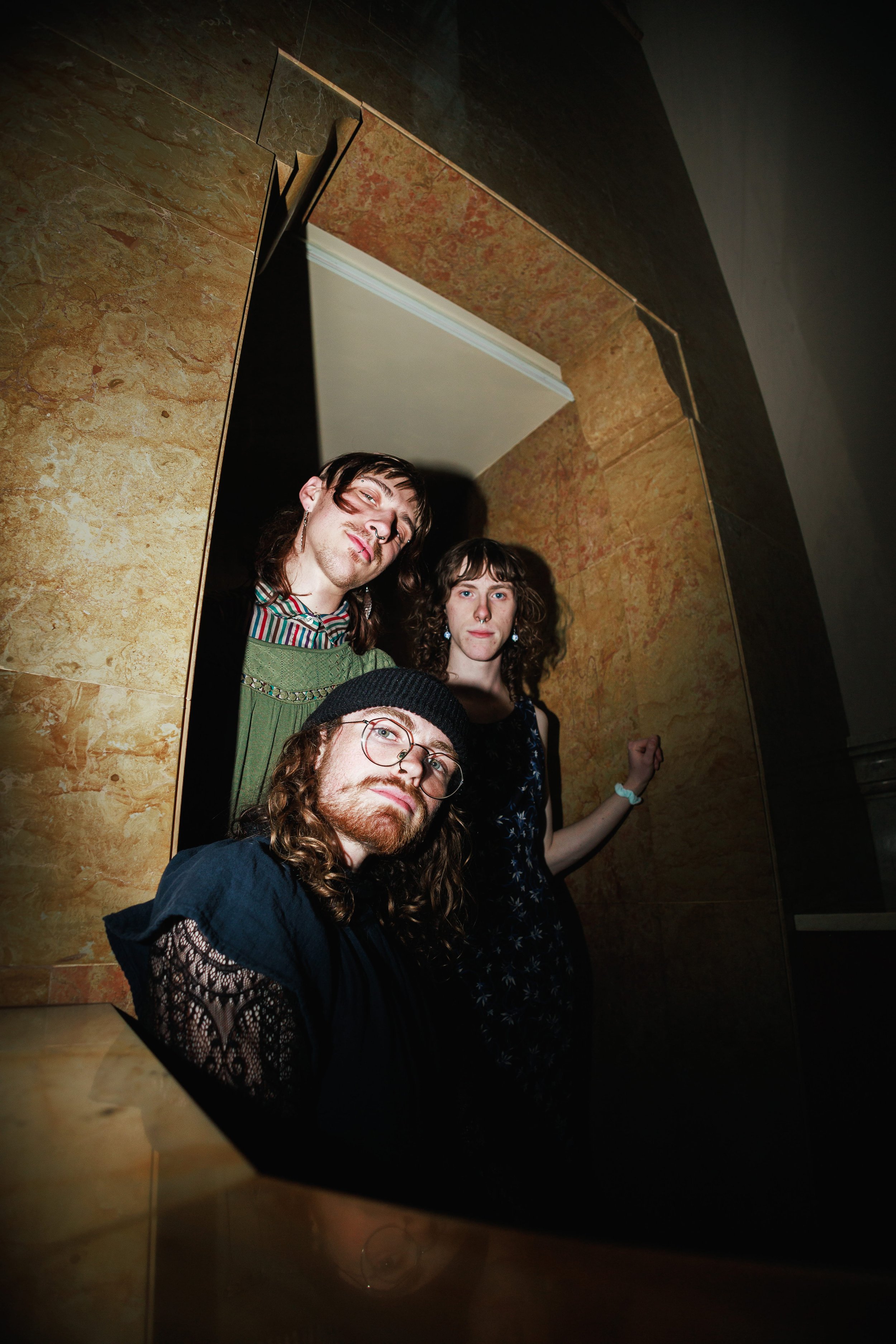A group of three people, two women and a man, standing in front of a mirror in a dimly lit room with tiled walls. The man has long hair, a beard, and glasses, and is looking directly at the camera. The woman in the middle has curly hair, wears earrin