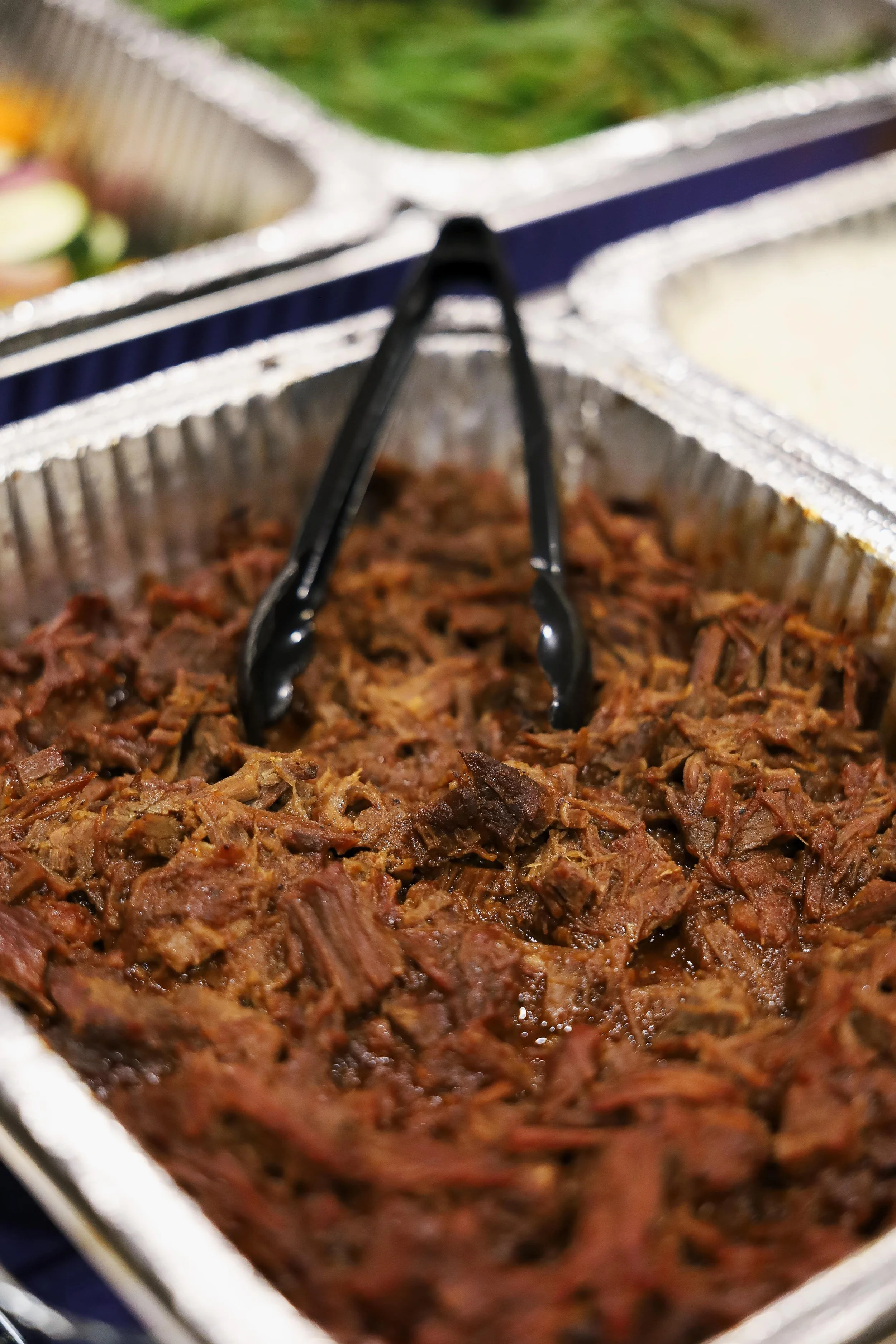 Shredded cooked beef in aluminum tray with black tongs