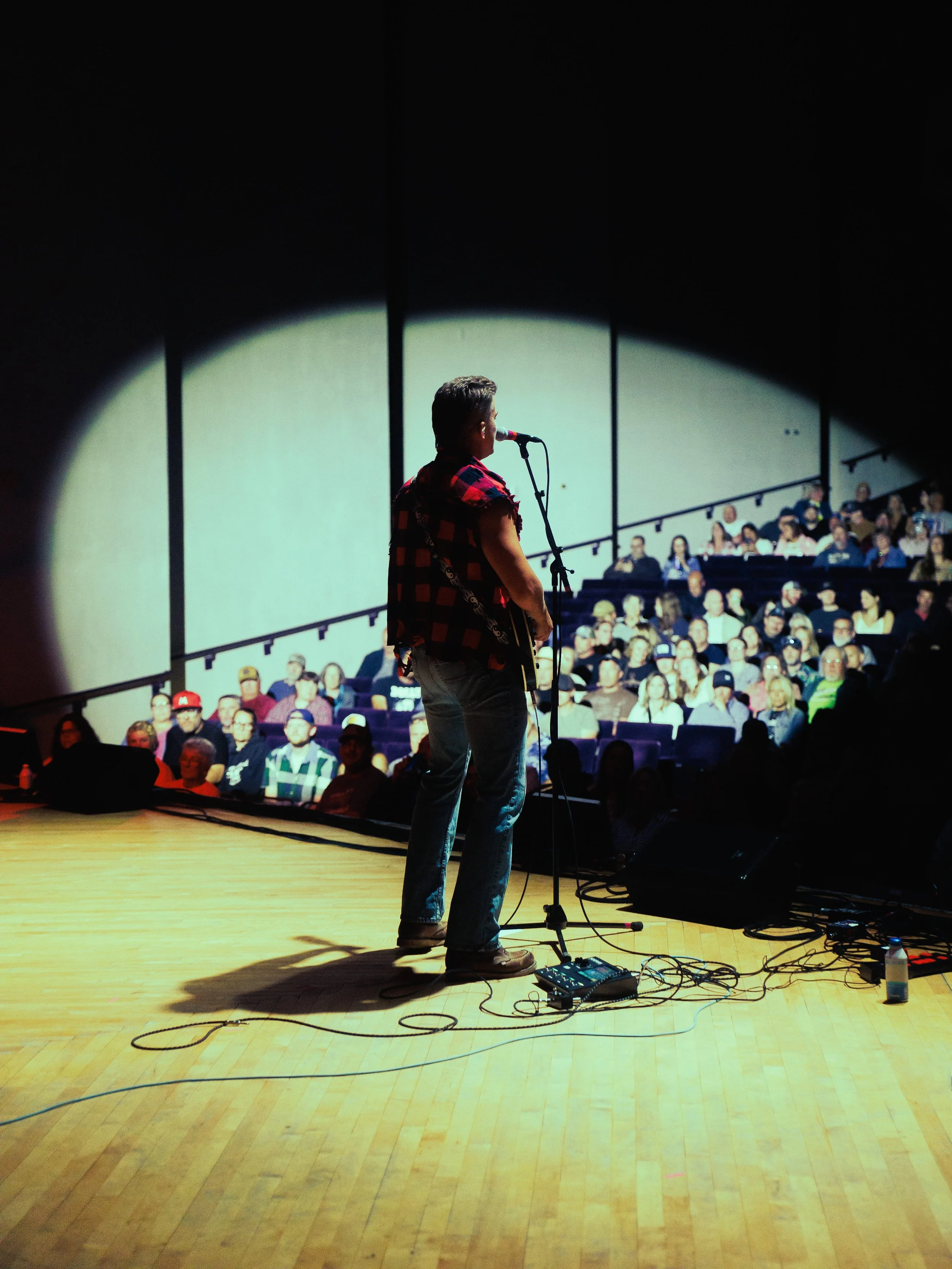 A man stands on a stage with a microphone, facing an audience in a dimly lit theater, with a spotlight on him and audio equipment on the floor.