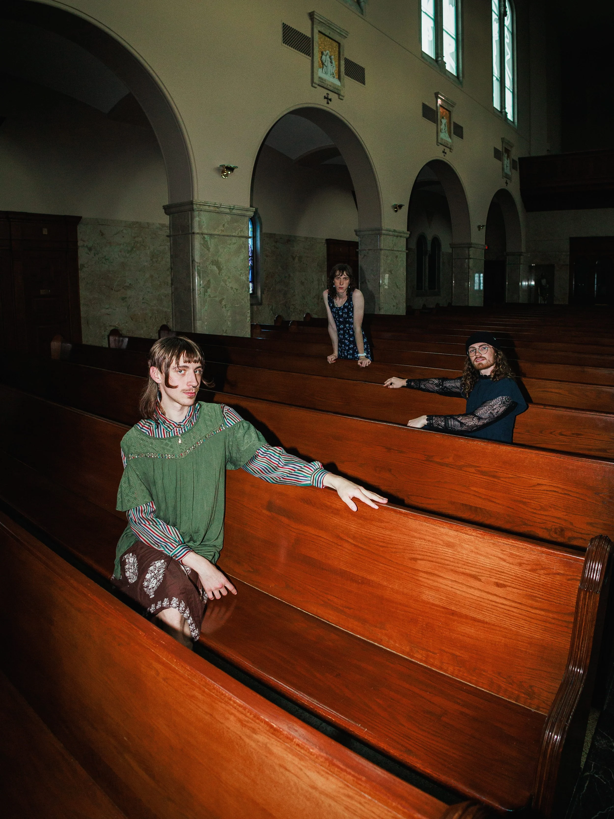 Three people sitting on wooden pews inside a church. A woman is leaning on a pew in the center, a man with long hair and a woman with glasses and curly hair are seated on the sides. The church has arched windows and religious images on the wall.