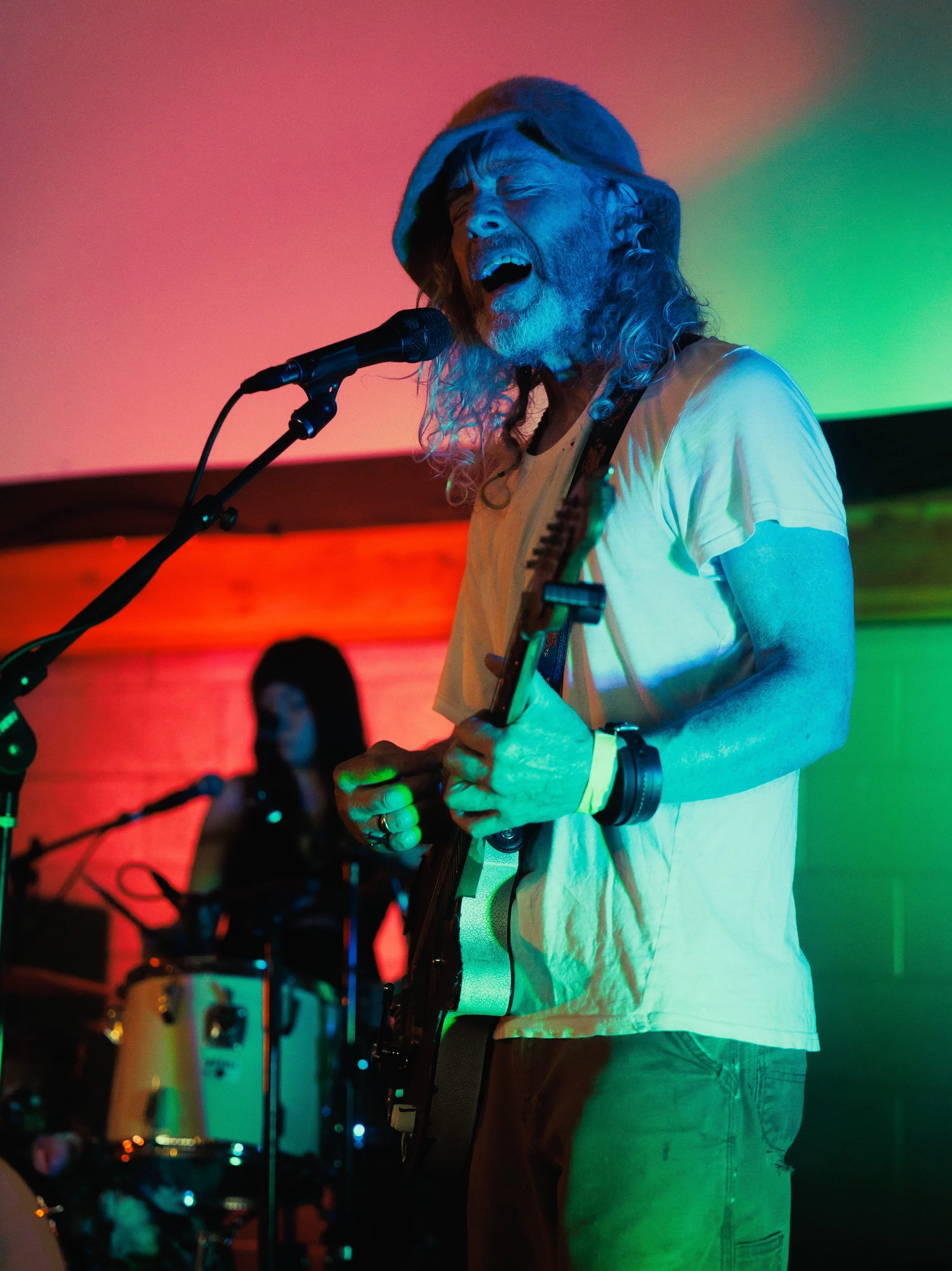 A musician singing passionately into a microphone while playing an acoustic guitar, wearing a hooded jacket, in colorful stage lighting with red, green, and yellow backgrounds.