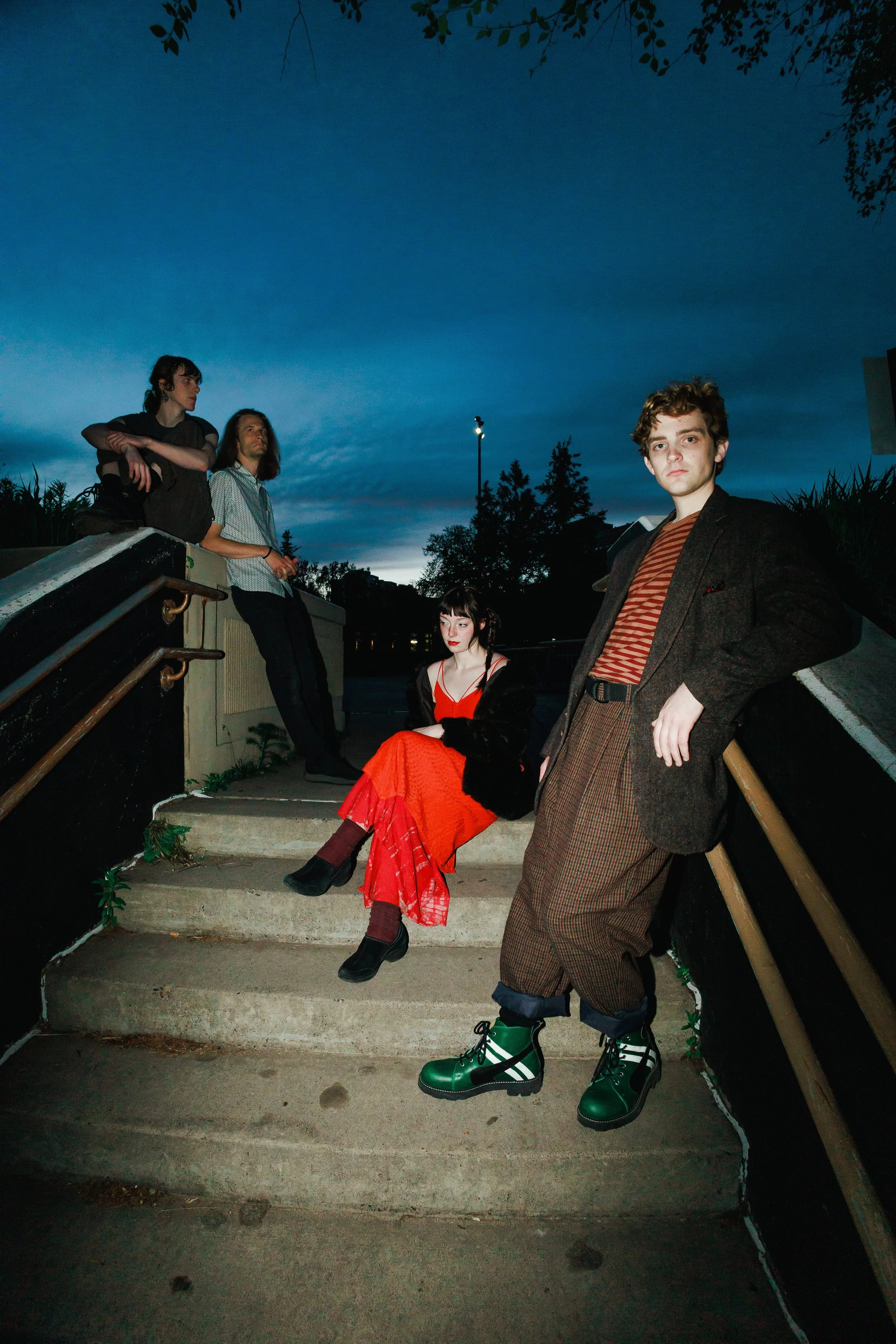 Group of five young adults on outdoor concrete steps at dusk, some sitting and some standing, with trees and a darkening sky behind them. The group has a casual and stylish appearance, with one individual wearing striking green and black boots.