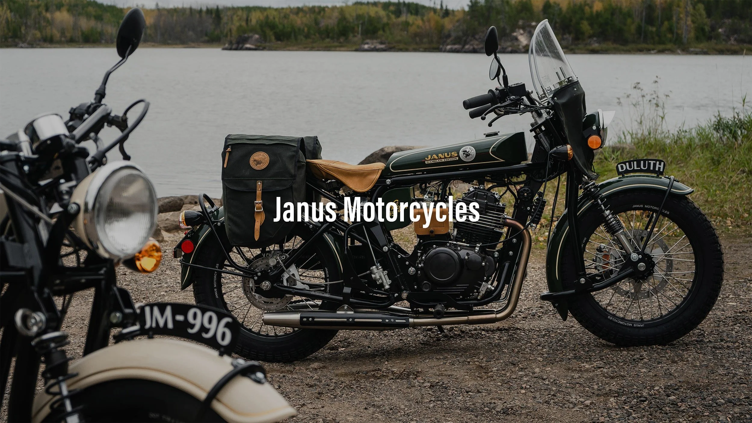 A vintage-style green motorcycle with a leather seat and a black saddlebag parked near a body of water with trees in the background.