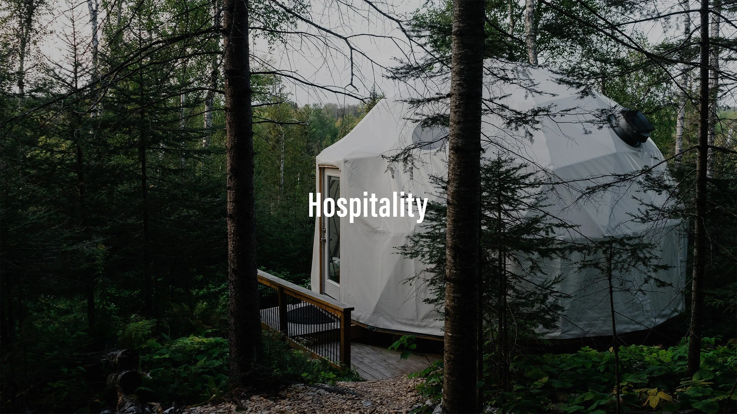 A geodesic dome-shaped tent in the woods with a small porch and glass door, surrounded by trees and greenery, labeled 'Hospitality'.
