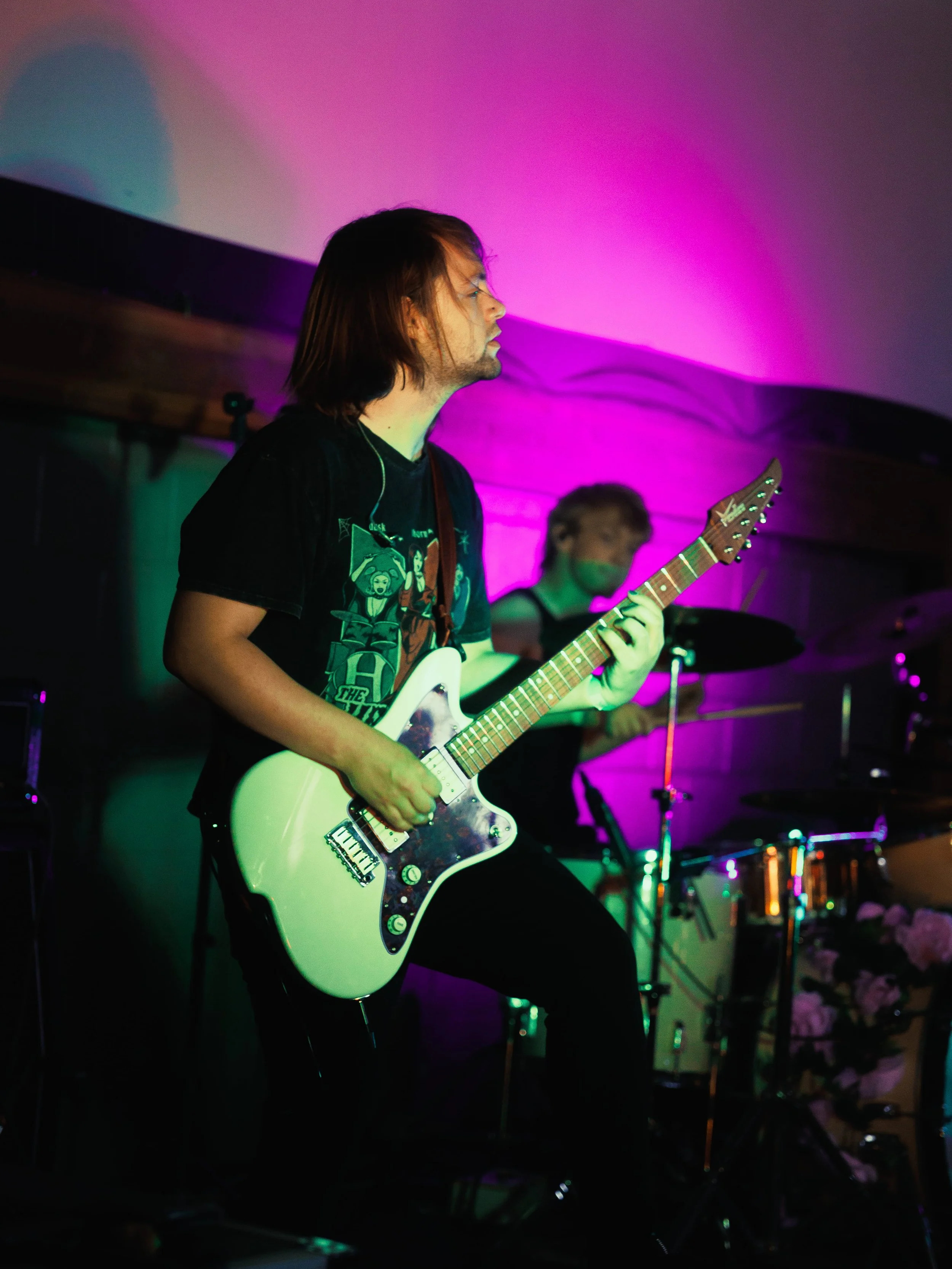 A person playing an electric guitar in a dimly lit room with colorful purple and green lighting, with a drummer visible in the background.