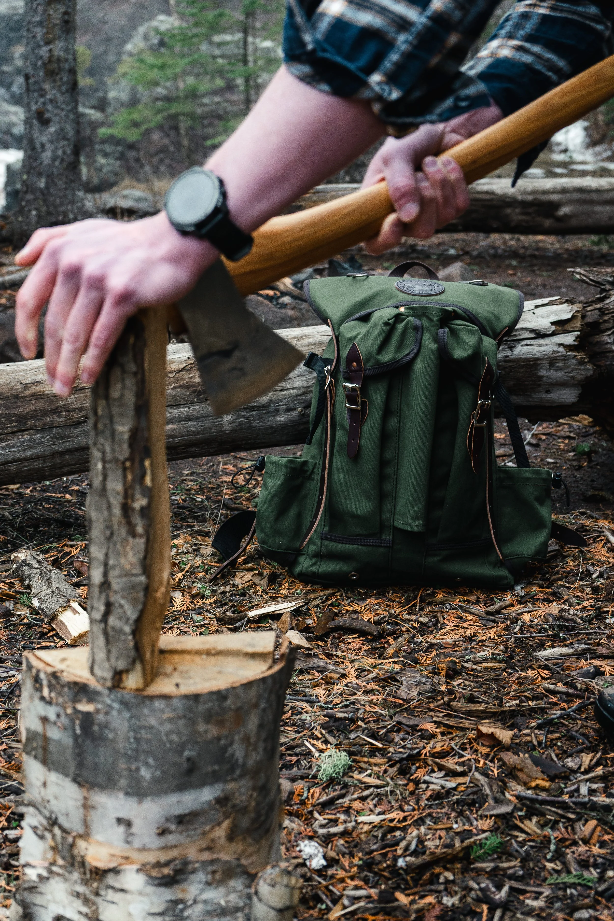 Close-up of a person chopping wood with an axe in a forest, with a green backpack on the ground nearby.