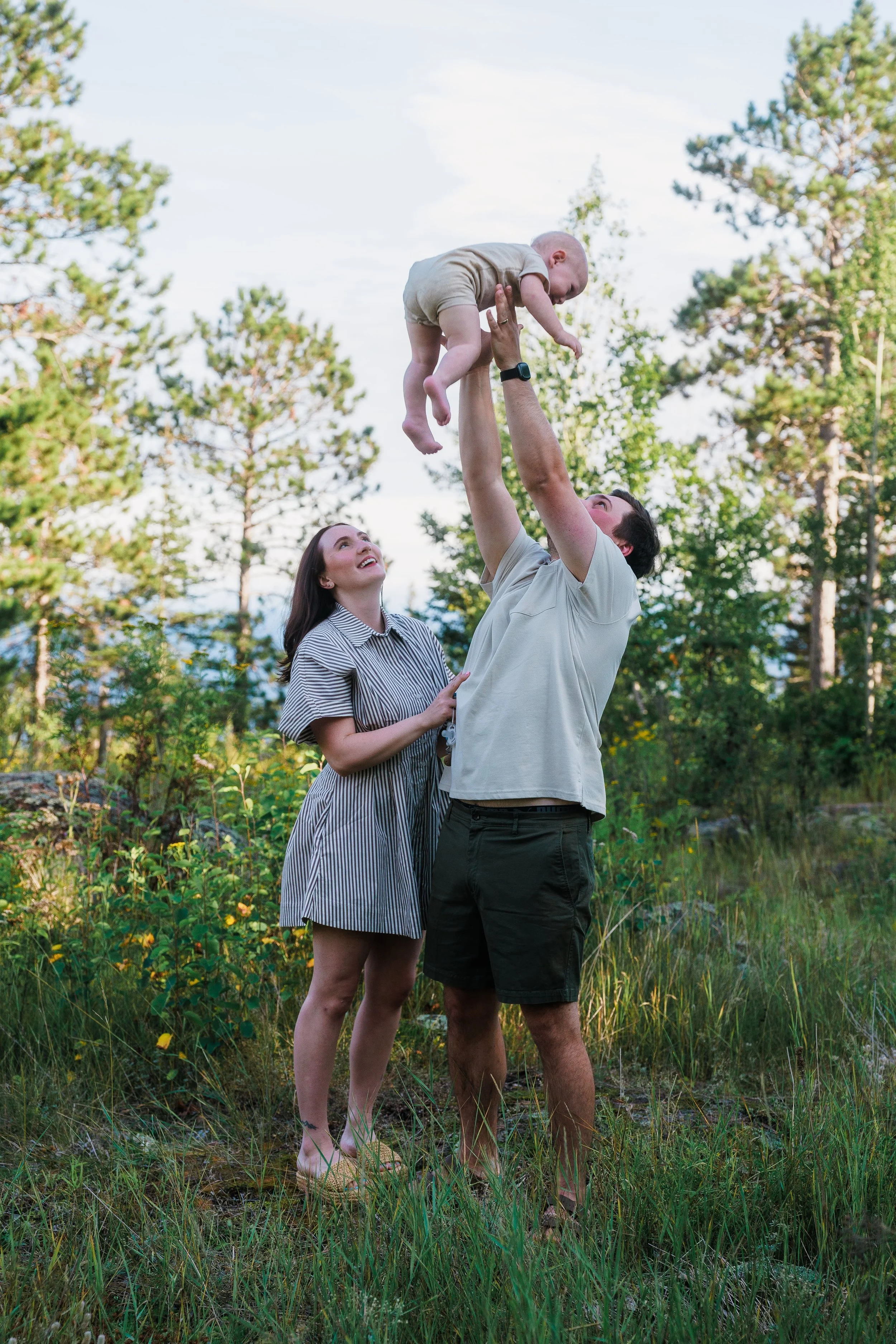 Family outdoors with a man lifting a baby and a woman watching in a grassy, wooded area.