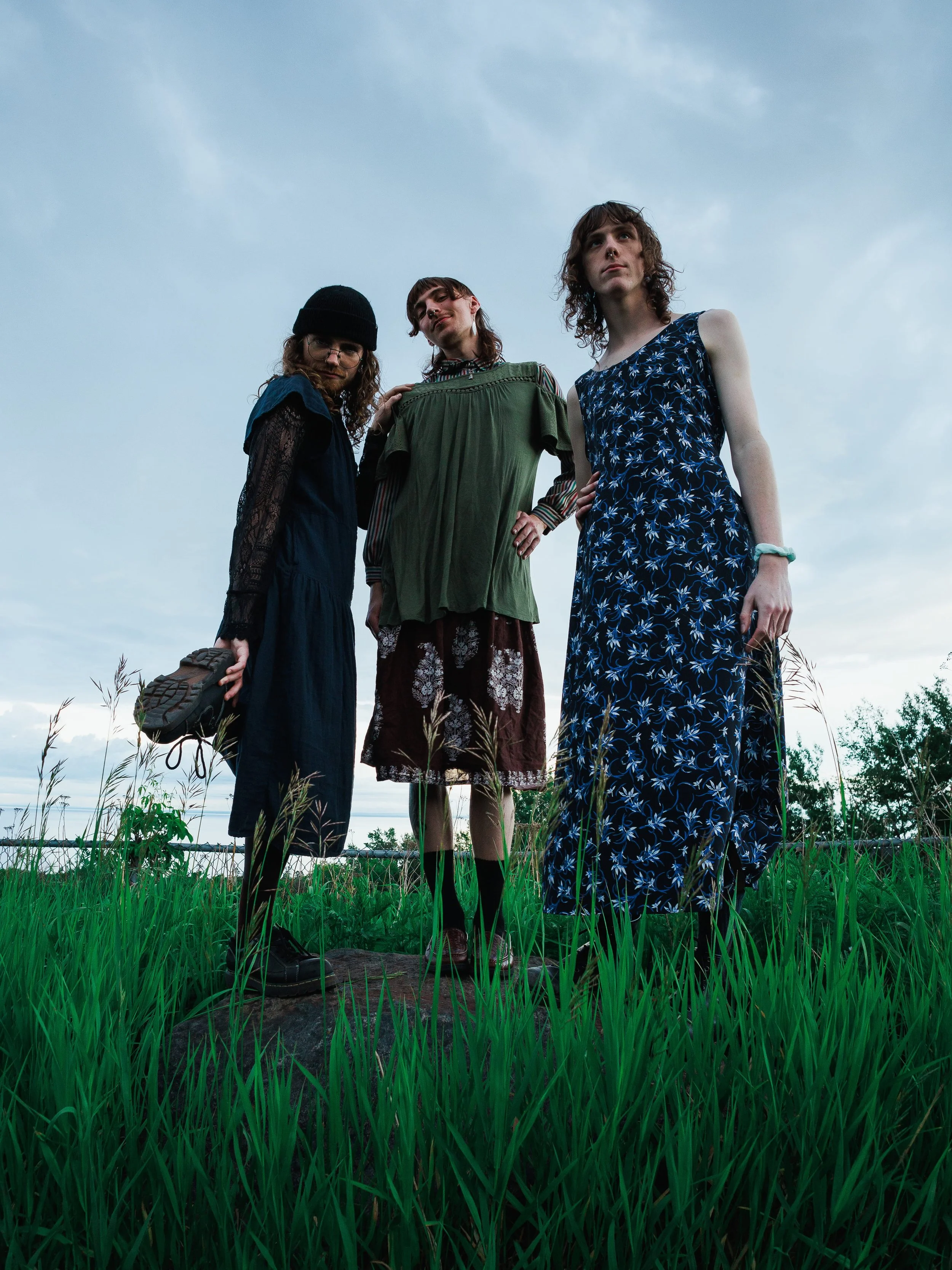 Three young women with curly hair standing outdoors in a grassy field, looking upwards against a partly cloudy sky.