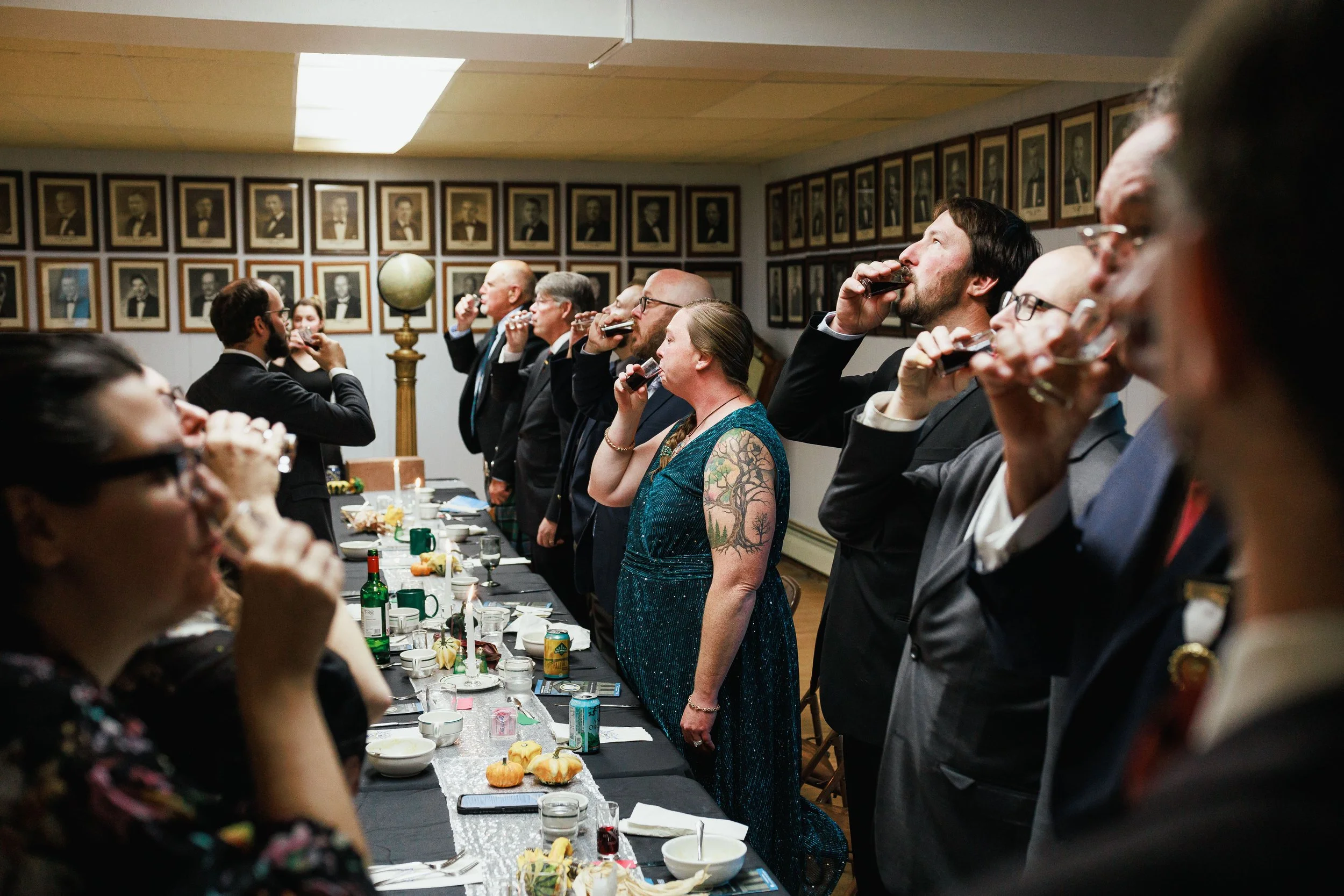 A group of people in formal attire standing around a table, drinking from shot glasses during a celebration event in a room with framed portraits on the walls.