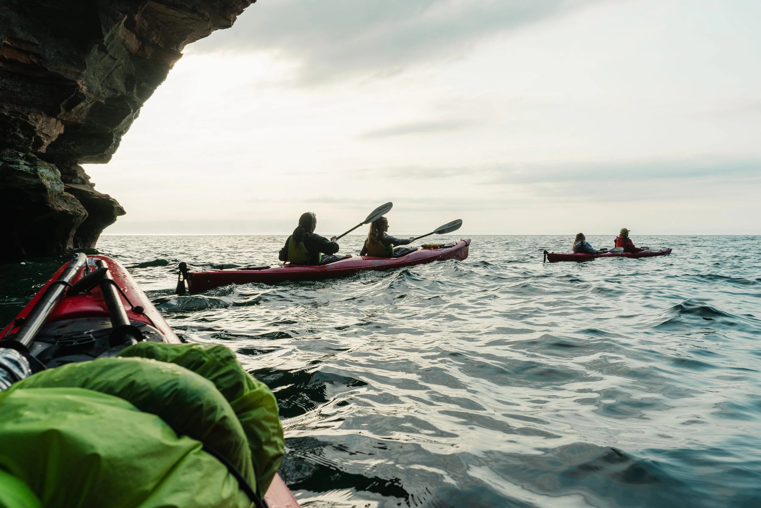 Kayakers paddling near a rocky cave along a body of water during overcast weather.
